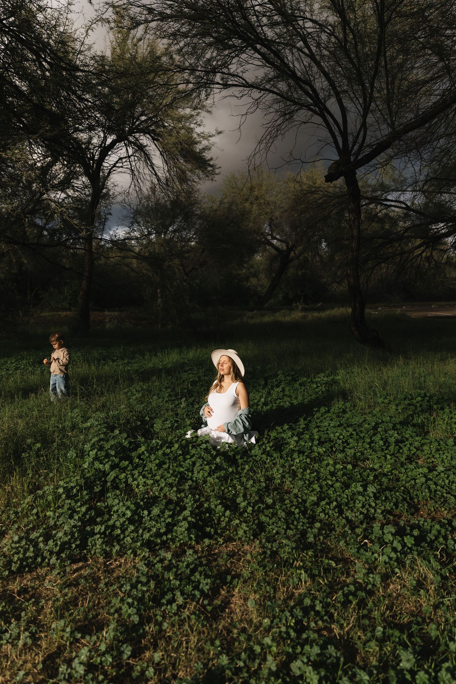 A person in white sits in a lush green field at sunset with trees silhouetted in the background.