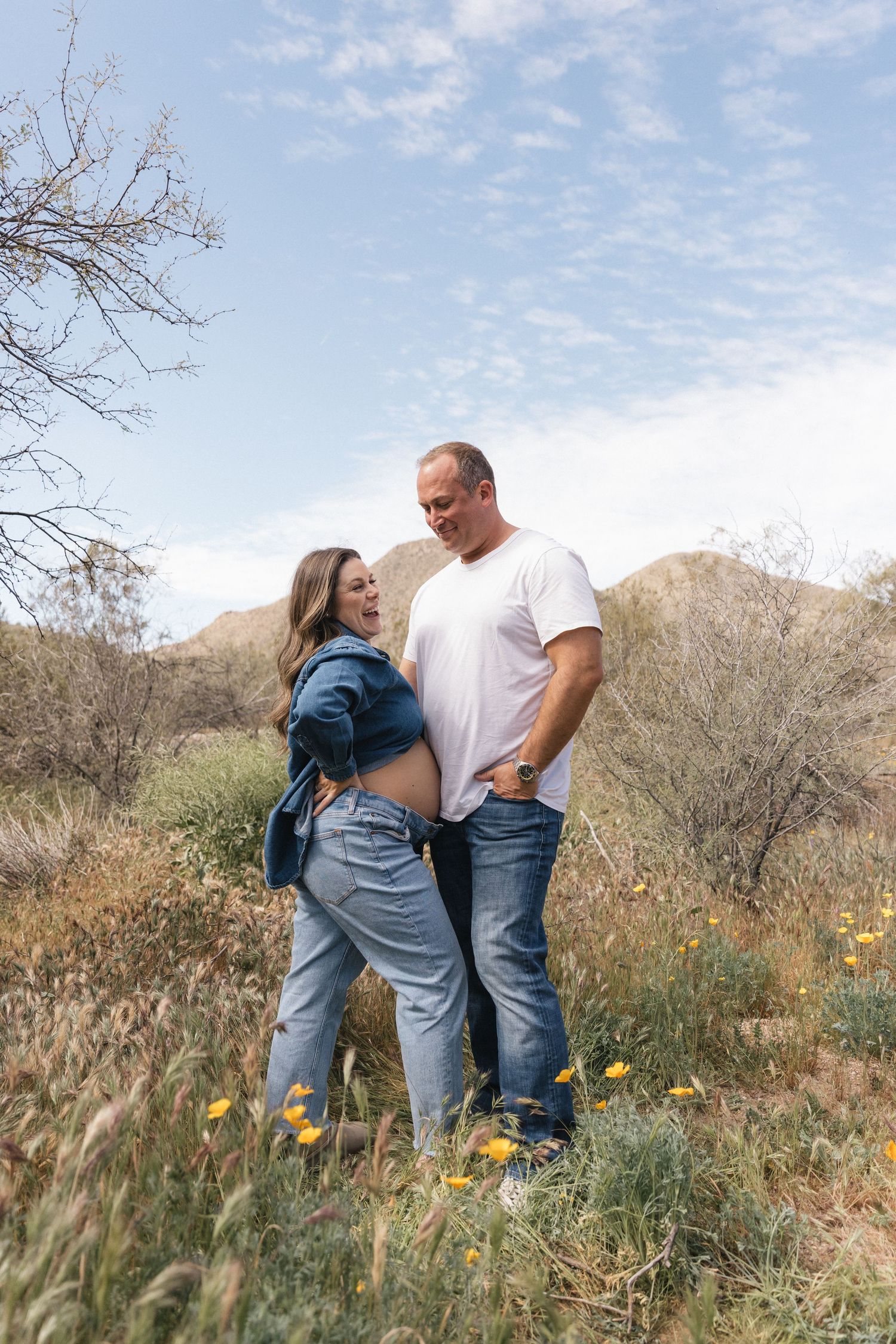 A couple stands together in a field of wildflowers with mountains and blue sky in the background.