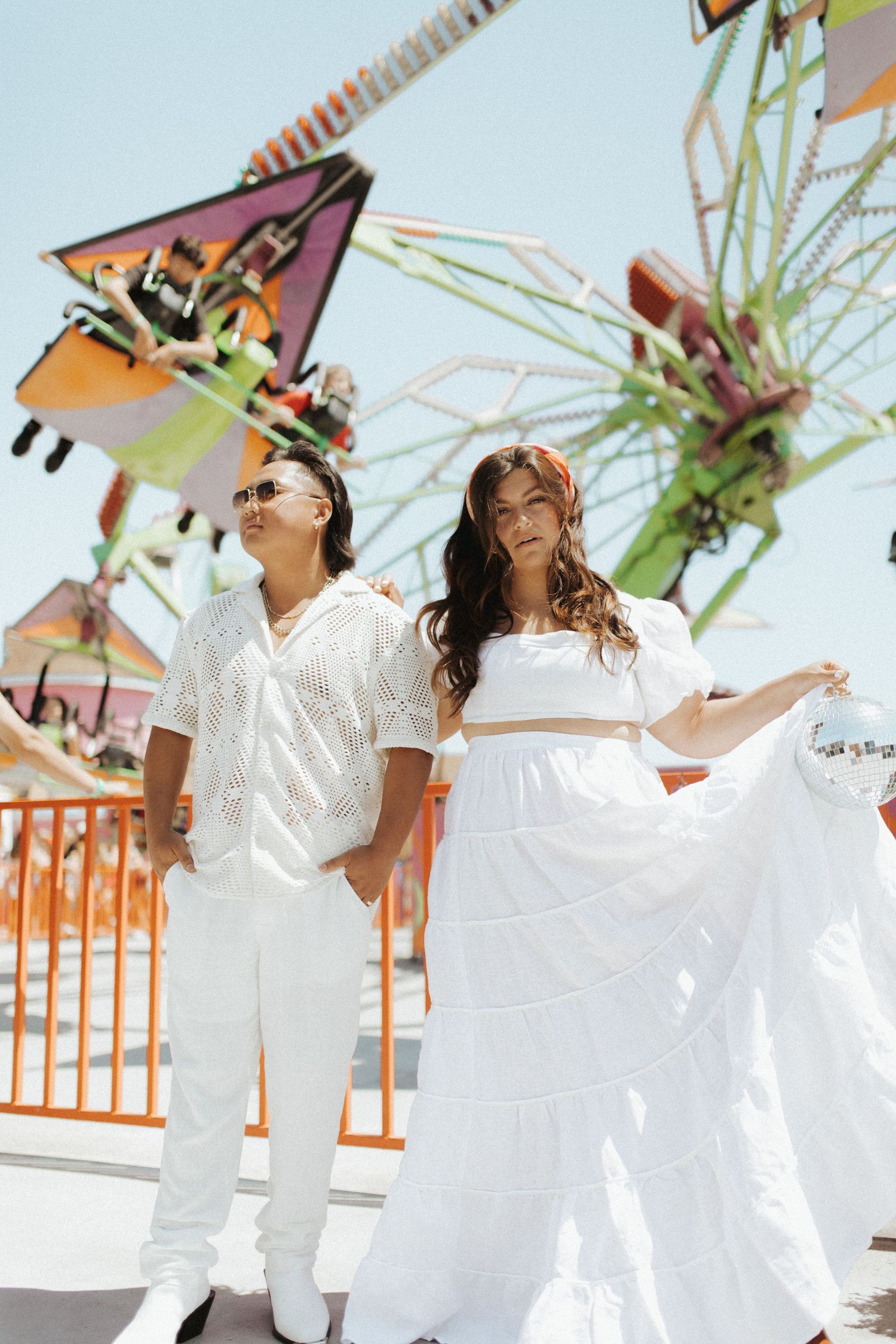 Couple in white outfits poses at carnival with colorful rides in background.