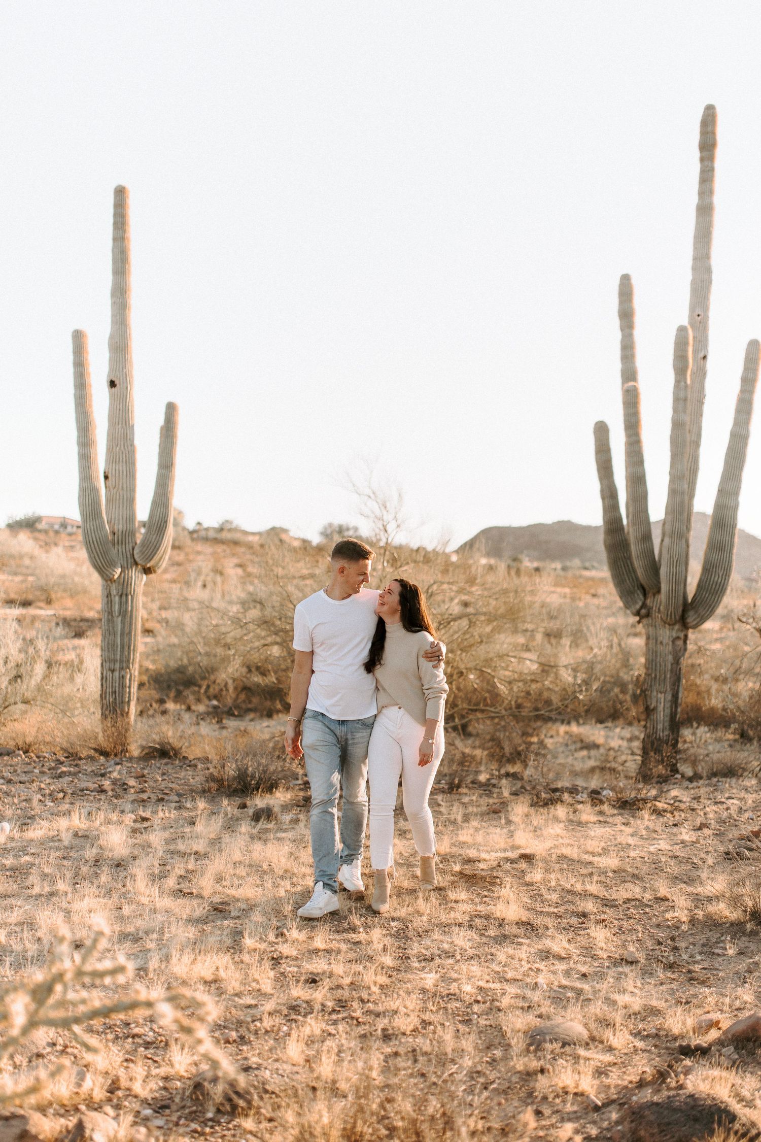 A couple walks together in a desert landscape with tall saguaro cacti at sunset.