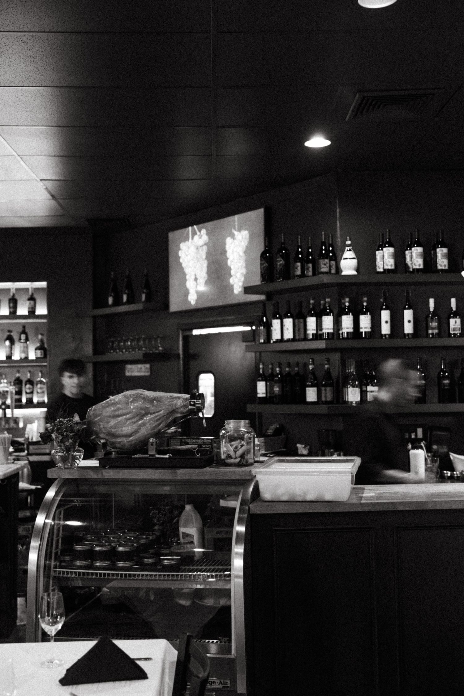 Black and white interior of a bar with bottles lined up on shelves and a display case.