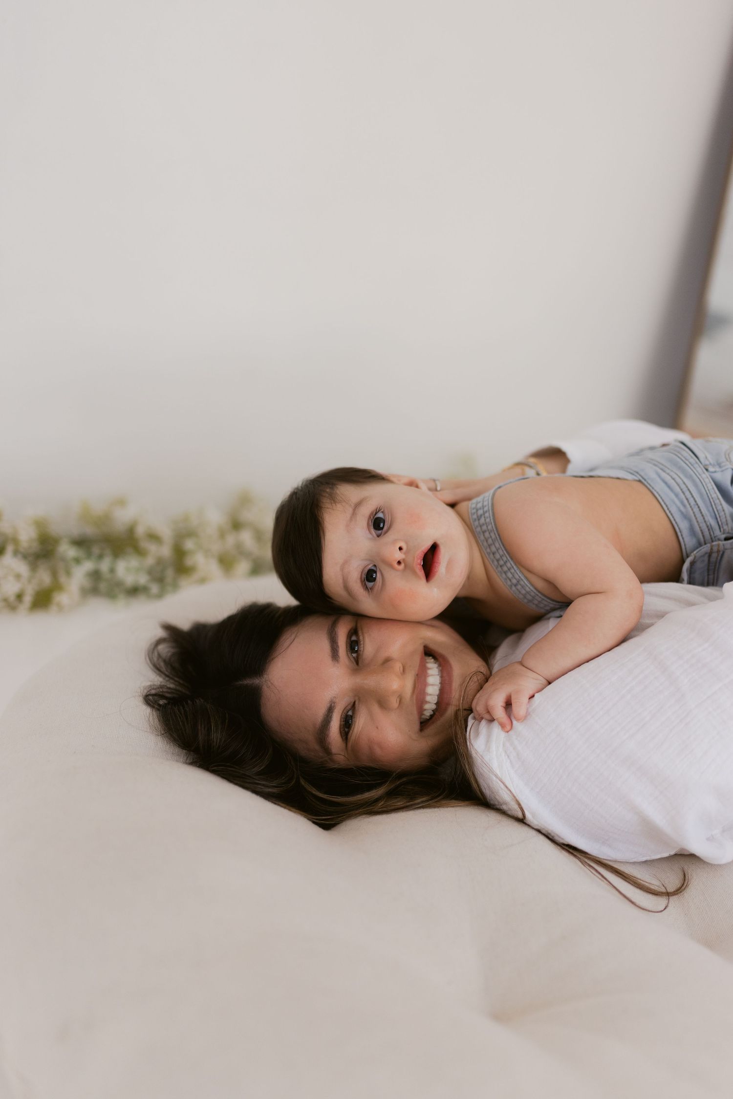 A smiling mother lies on a bed with her happy baby resting on her back, sharing a joyful moment together.