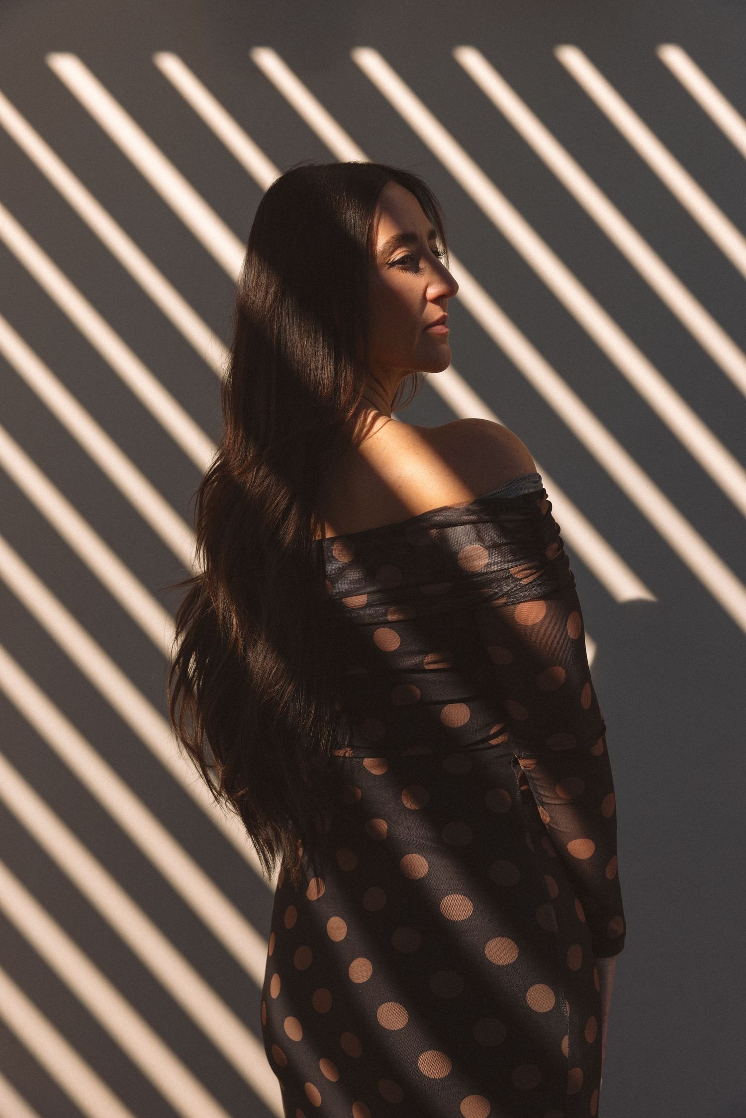 Woman in polka dot off-shoulder dress with long hair, posed against wall with diagonal light and shadow stripes.
