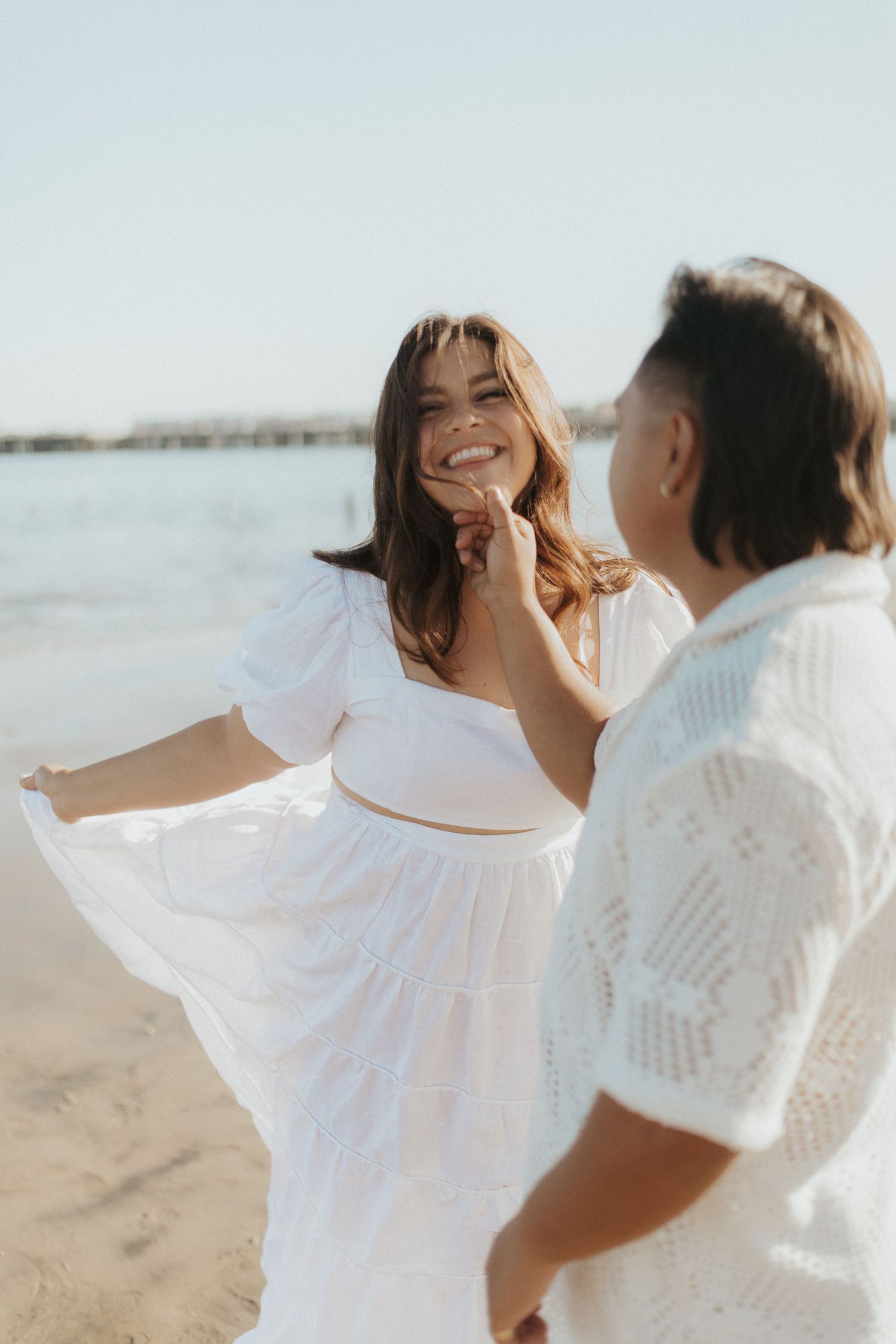 Couple in white outfits dancing and laughing together on sunny beach.