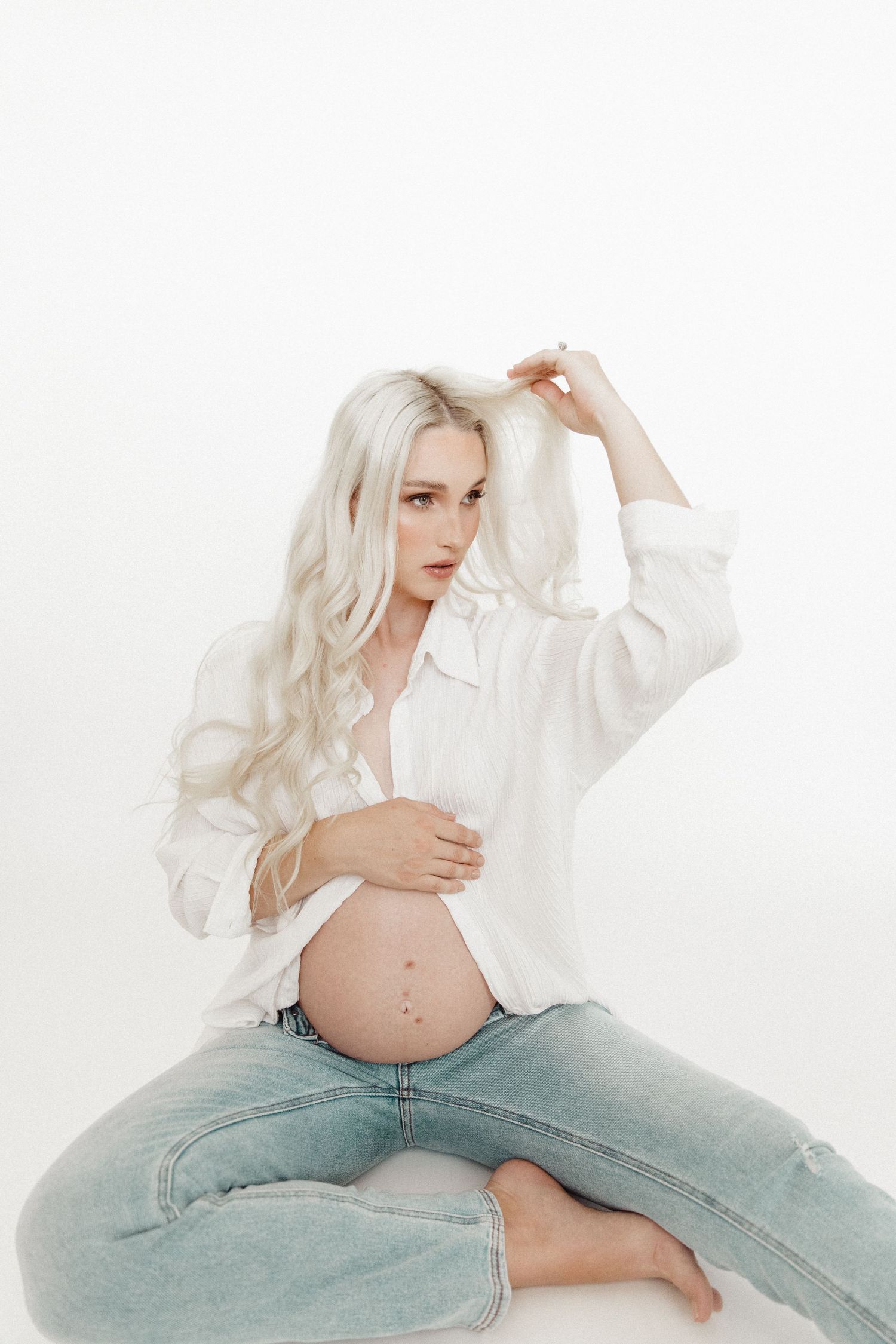 Artistic maternity portrait in white shirt and jeans against white background.