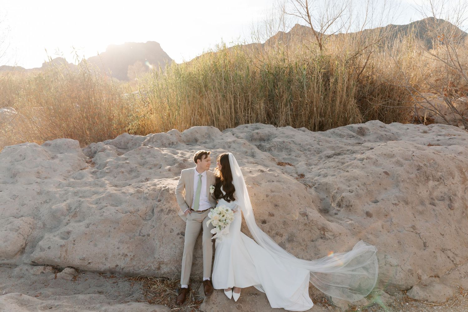 Couple in wedding attire poses against scenic desert landscape with mountains and sunset in background.