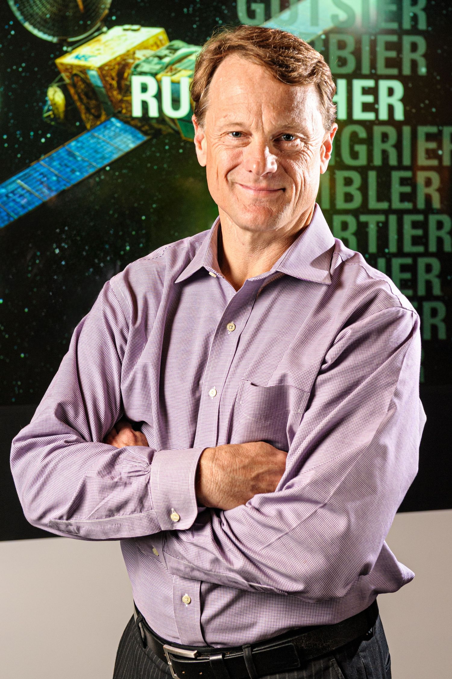 Aerospace executive poses in purple shirt against satellite backdrop.