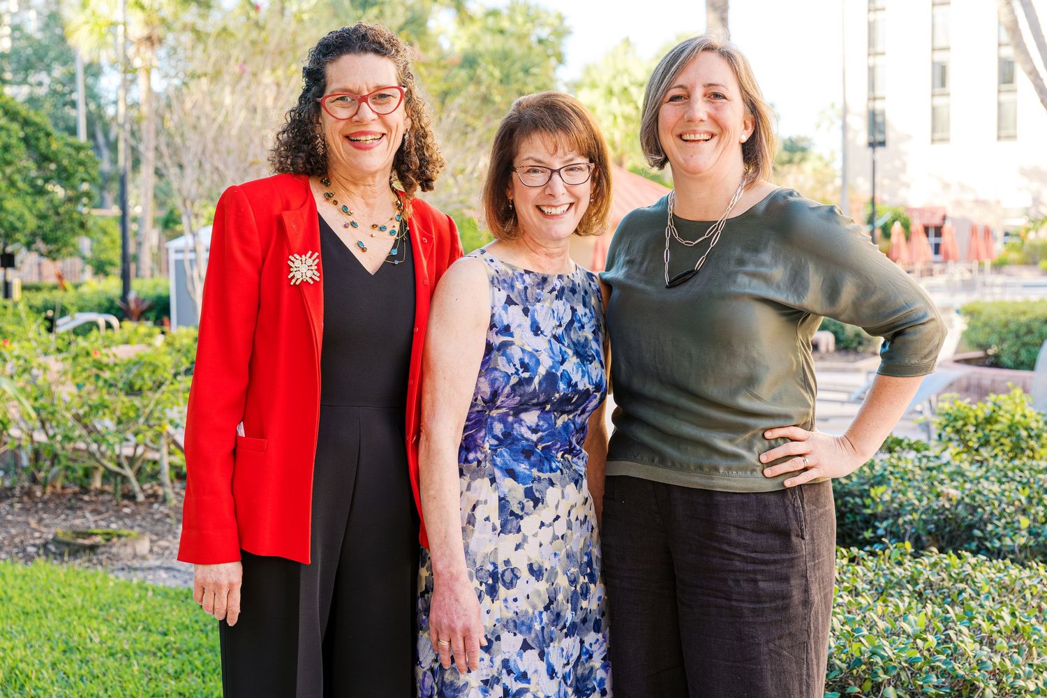 Three people stand together outdoors on a sunny day wearing colorful formal attire.
