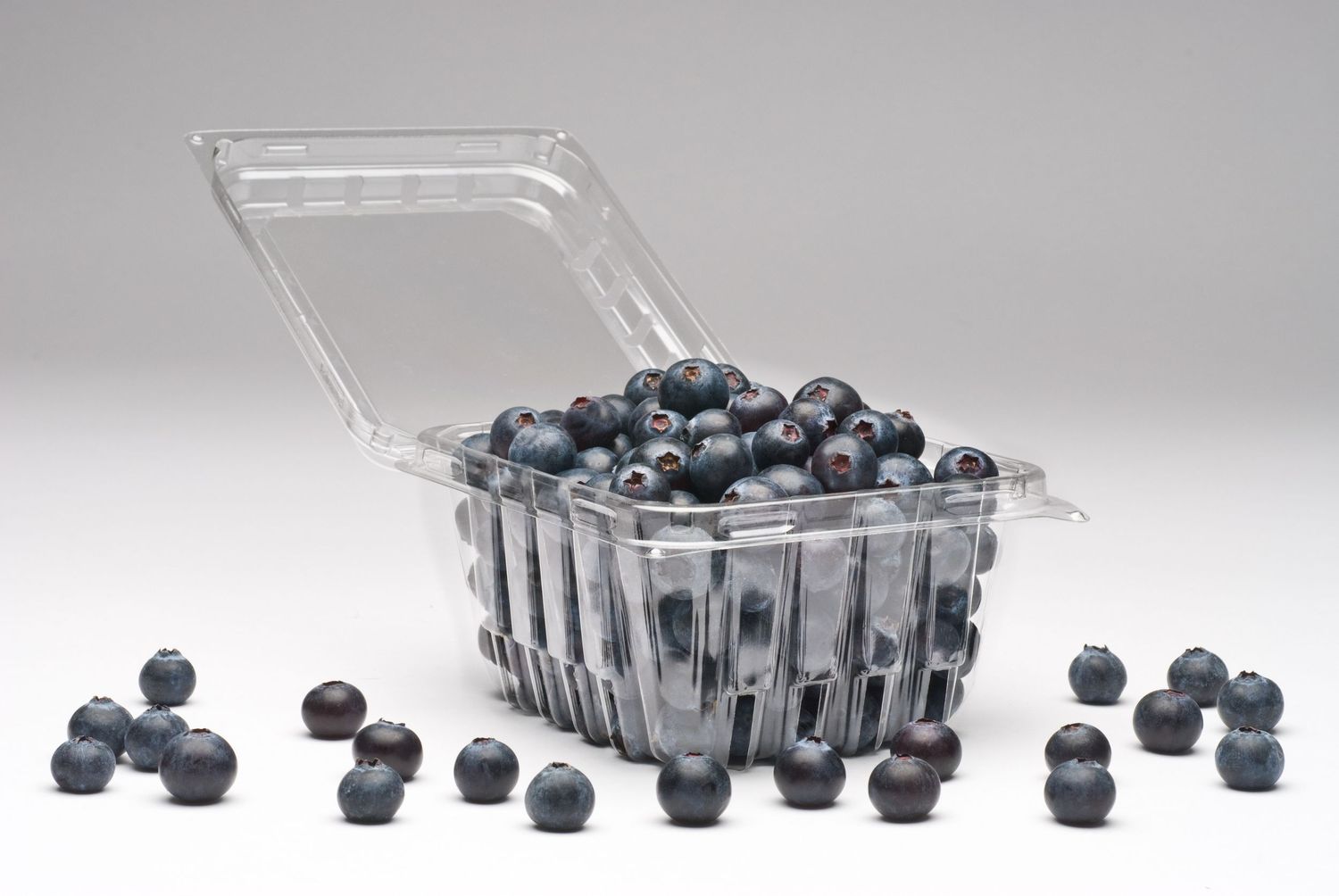 Ripe blueberries scattered around a clear plastic produce container on a white surface.