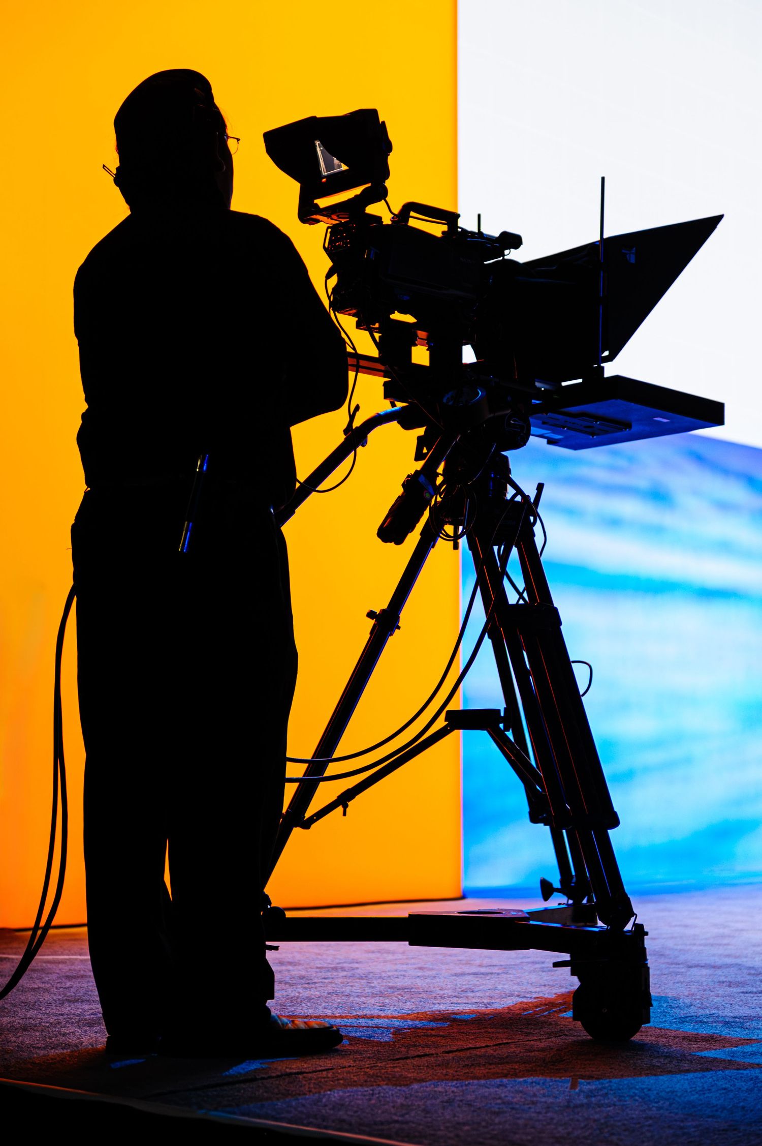 Silhouette of camera operator with professional video equipment against orange and blue background.