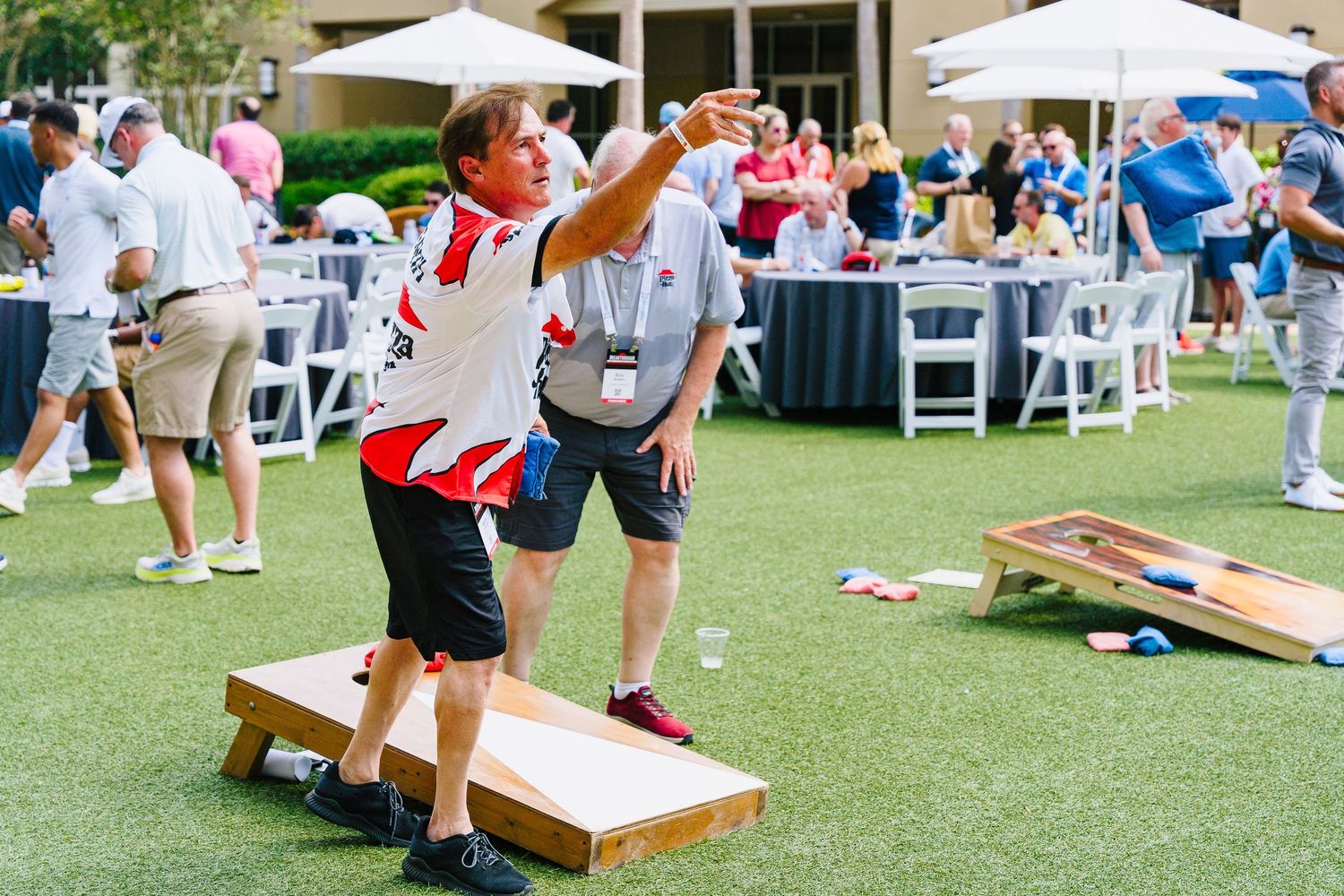 Players competing in outdoor cornhole tournament on green lawn with tables in background.