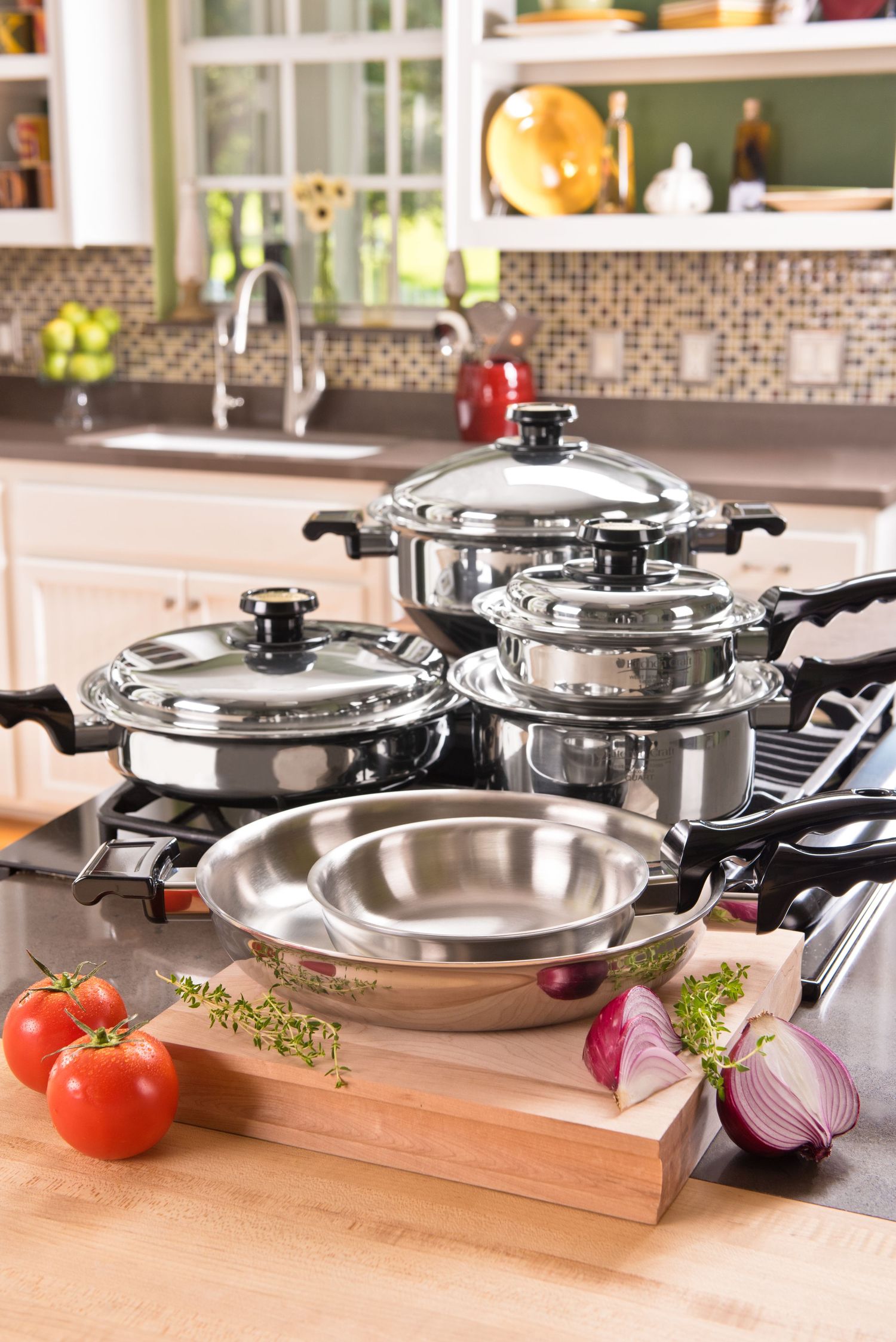 Stainless steel cookware set with pots and pans displayed on stovetop next to fresh vegetables on wooden cutting board.