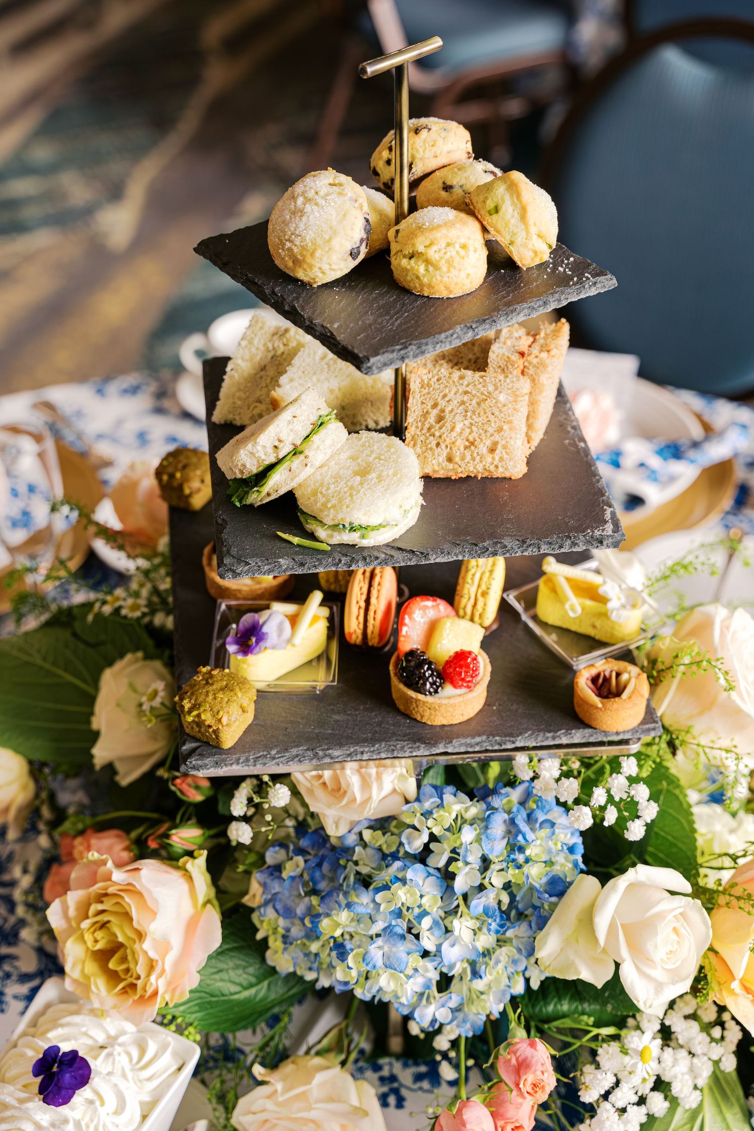 Elegant afternoon tea service with mini sandwiches and pastries displayed on tiered stands alongside blue hydrangeas and roses.