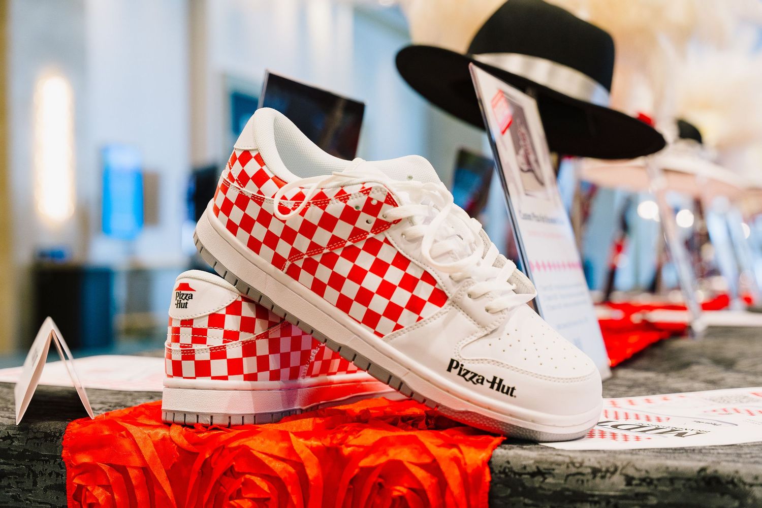 Red and white checkered sneakers displayed on red platform with black hat.