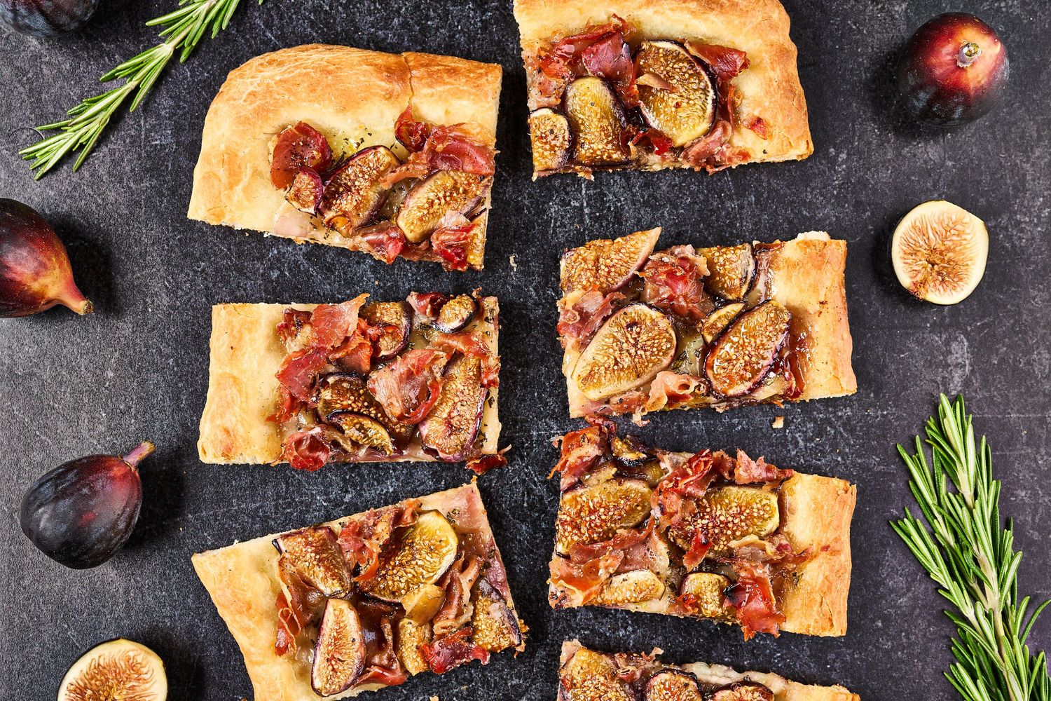 Rustic flatbread slices topped with fresh figs, herbs, and balsamic glaze on dark stone surface with rosemary sprigs.