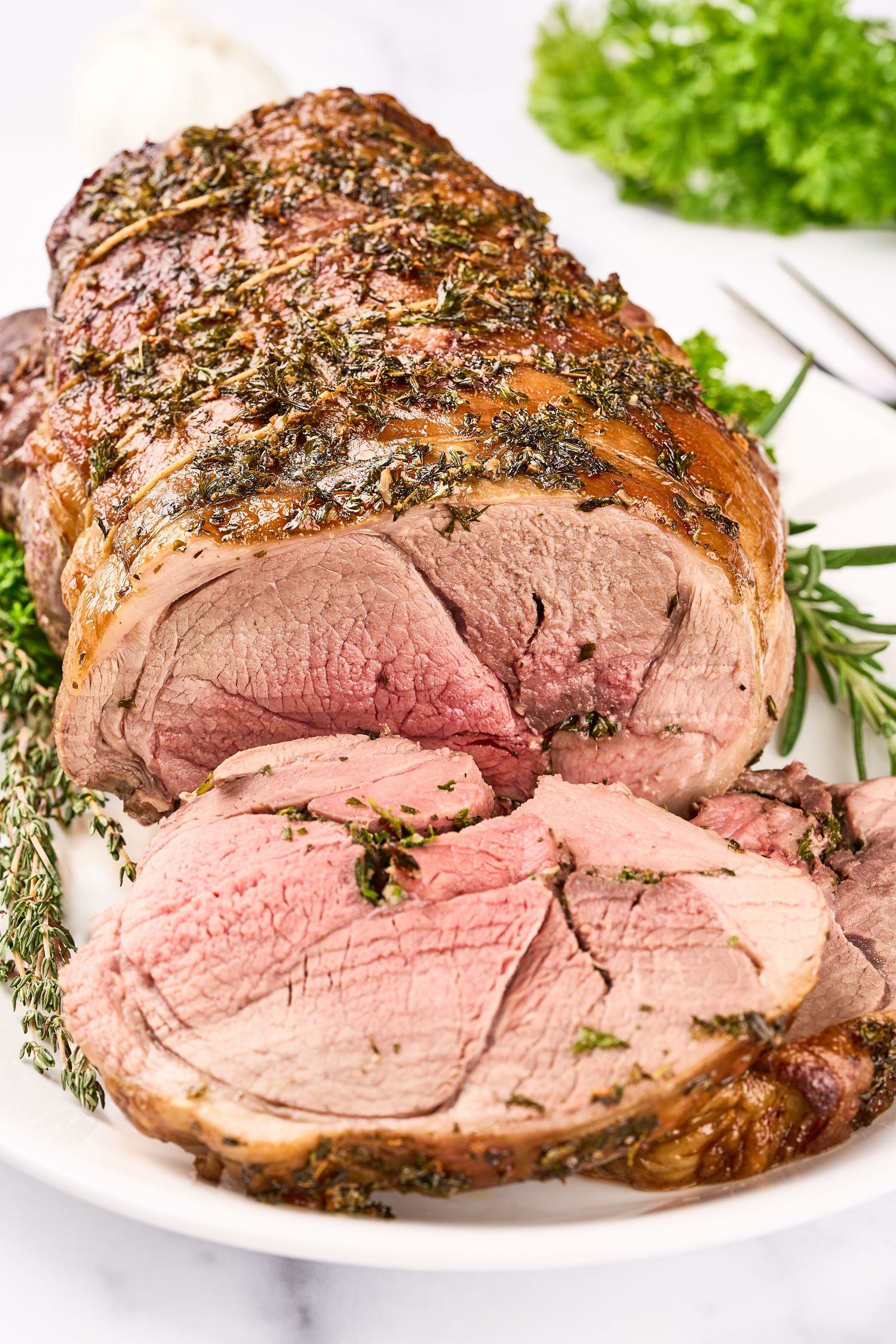 Sliced herb-crusted roasted leg of lamb cooked medium rare and served with fresh rosemary garnish on white plate.