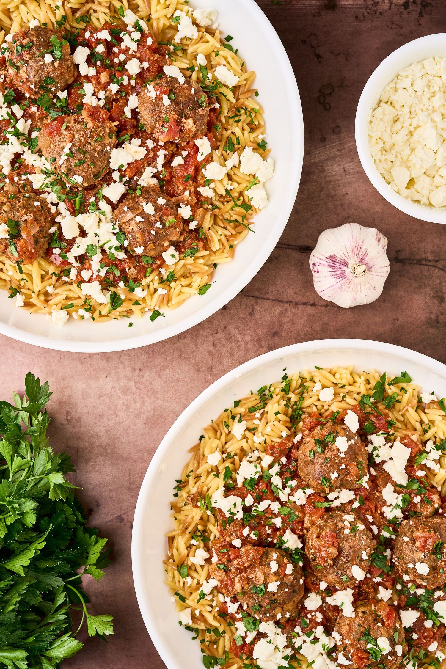 Two plates of Middle Eastern orzo pasta topped with meatballs, feta cheese and fresh herbs served with garlic on wooden table.