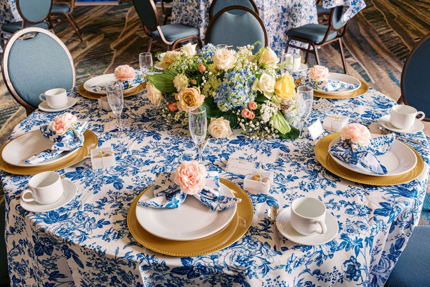 Elegant table settings with blue and white floral tablecloth, gold charger plates, and delicate floral centerpieces.
