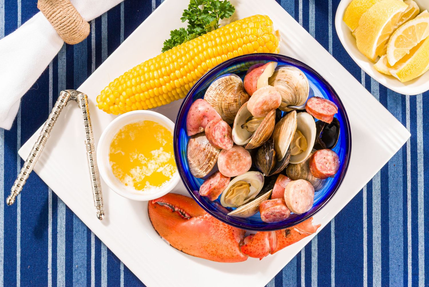 Assorted colorful food dishes including corn on the cob, shrimp, and soups displayed on blue and white striped background.