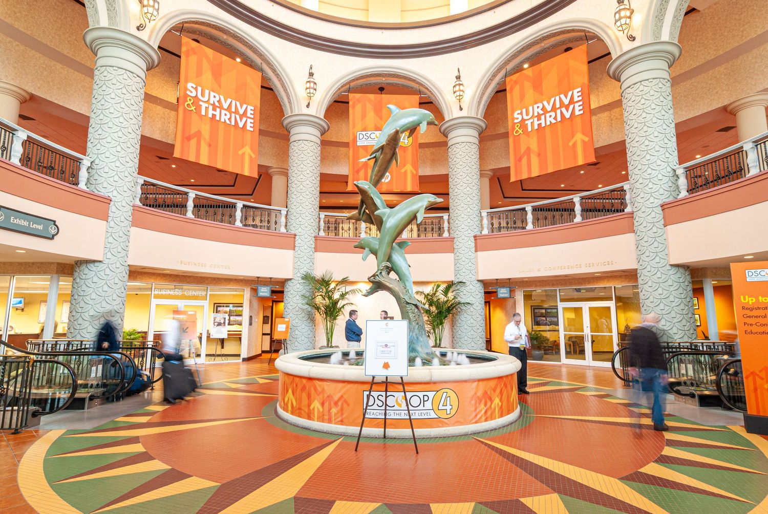 Modern shopping mall interior with ornate columns and a central fountain display beneath orange banners.