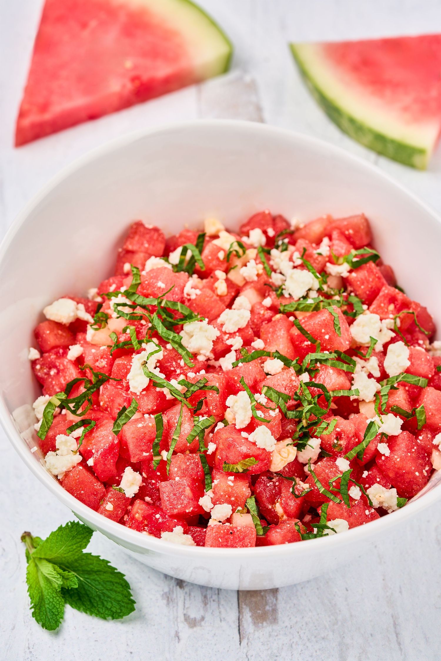 Fresh watermelon salad with crumbled feta cheese, mint, and herbs served in a white bowl on light surface.