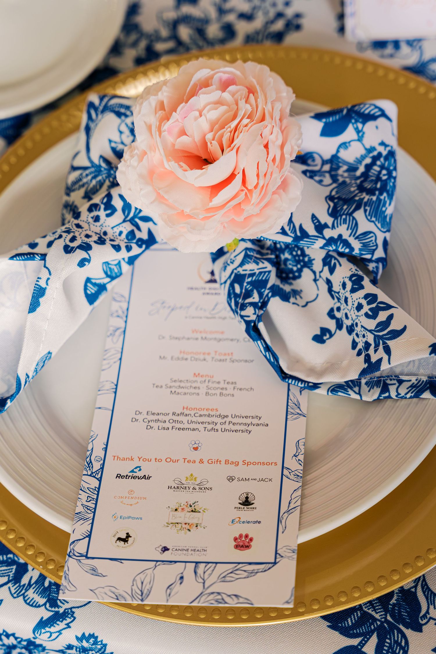 Wedding invitation with blue floral border design and pink rose accent displayed on gold-rimmed plates.