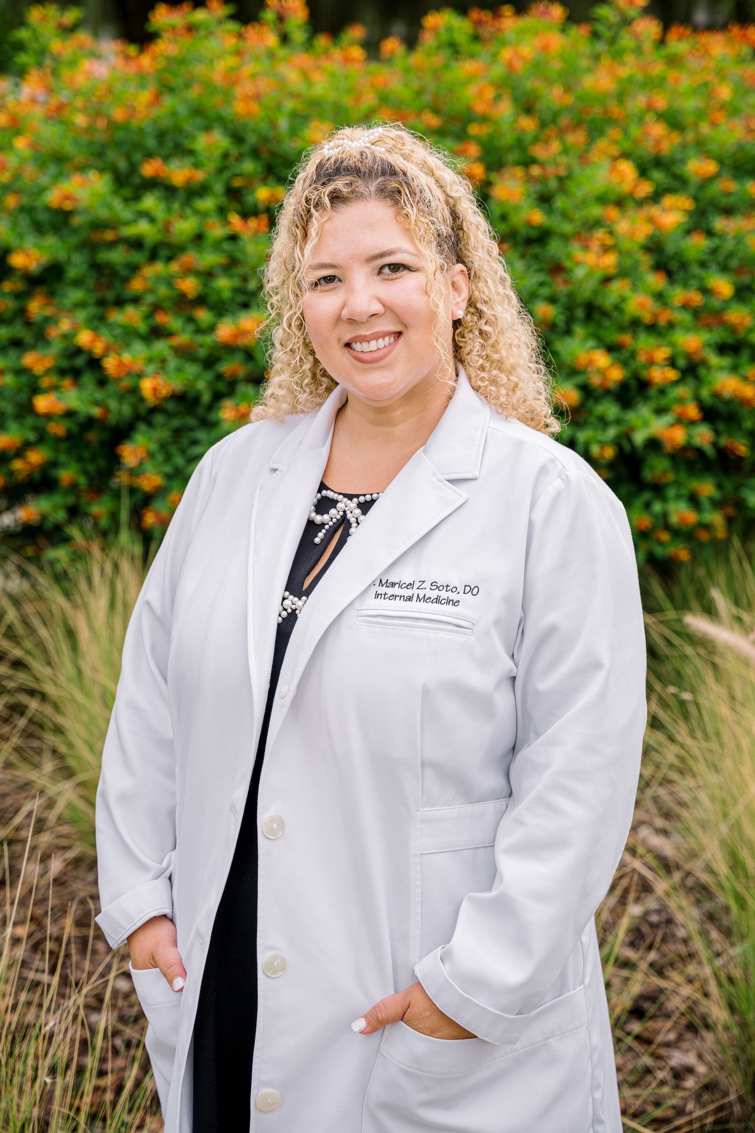 Healthcare professional in white coat standing outdoors with orange flowers in background.