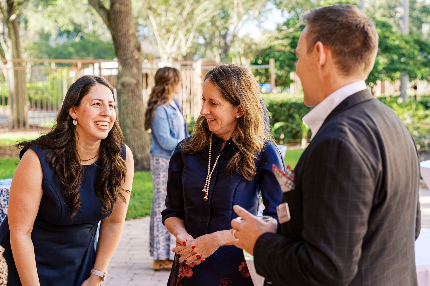 People in formal attire gather outdoors for a social event, laughing and conversing in a garden setting.