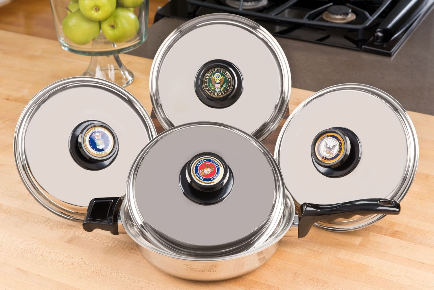 Four stainless steel pot lids with black knobs arranged in a diamond pattern on a wooden kitchen counter.
