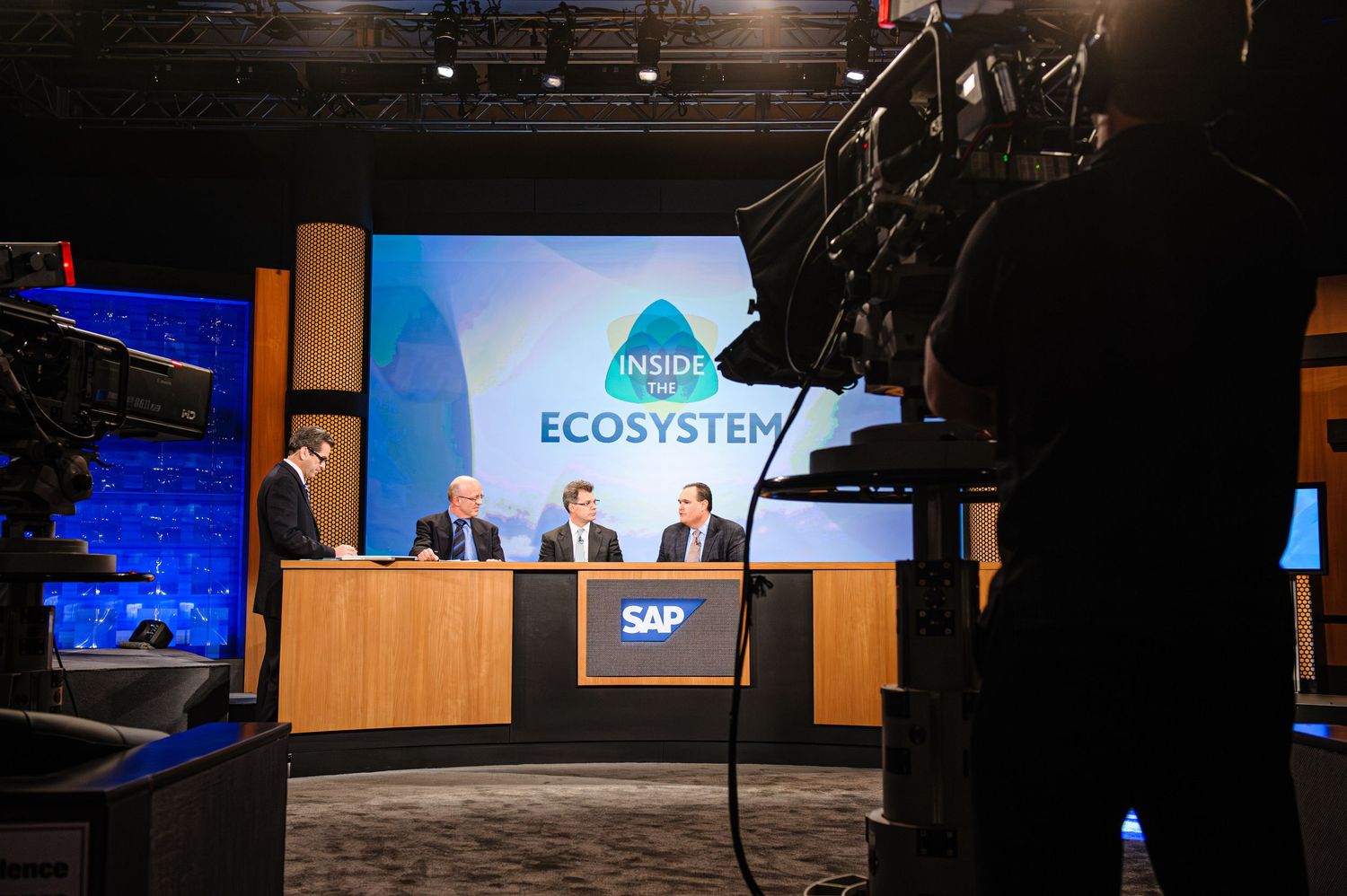 Behind-the-scenes view of television broadcast studio with Ecosystem branding on stage.