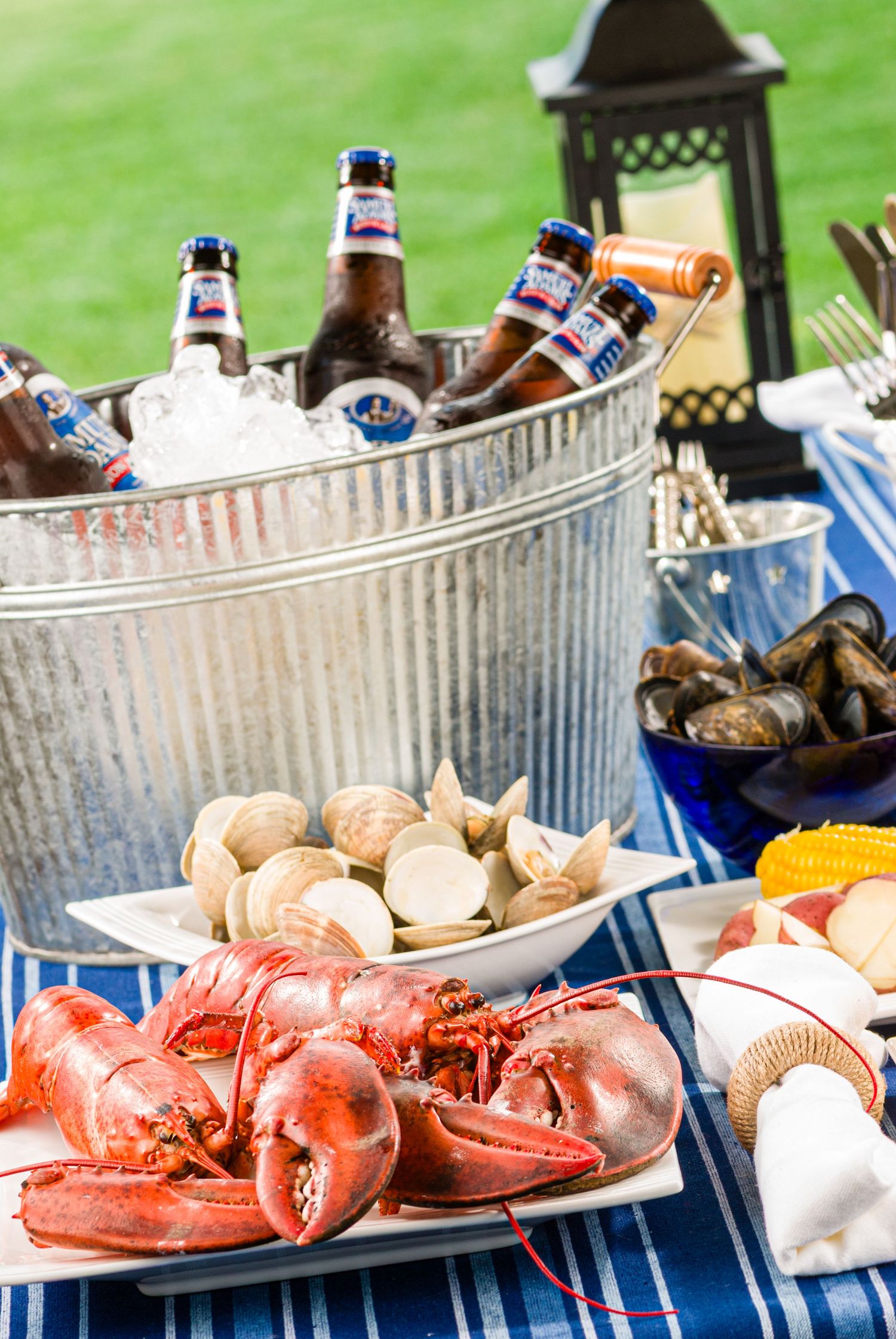 Cold beer bottles chill in a metal tub alongside fresh seafood and cooked lobsters on a blue tablecloth at an outdoor party.