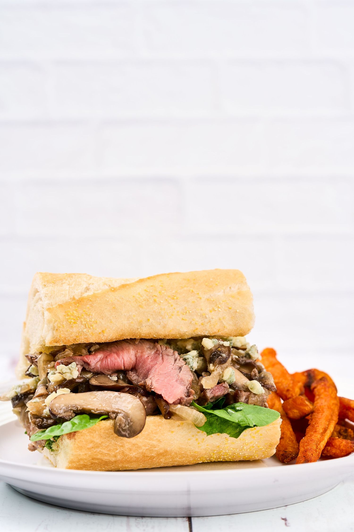 Juicy steak sandwich with mushrooms and greens served on a white plate with sweet potato fries on the side.