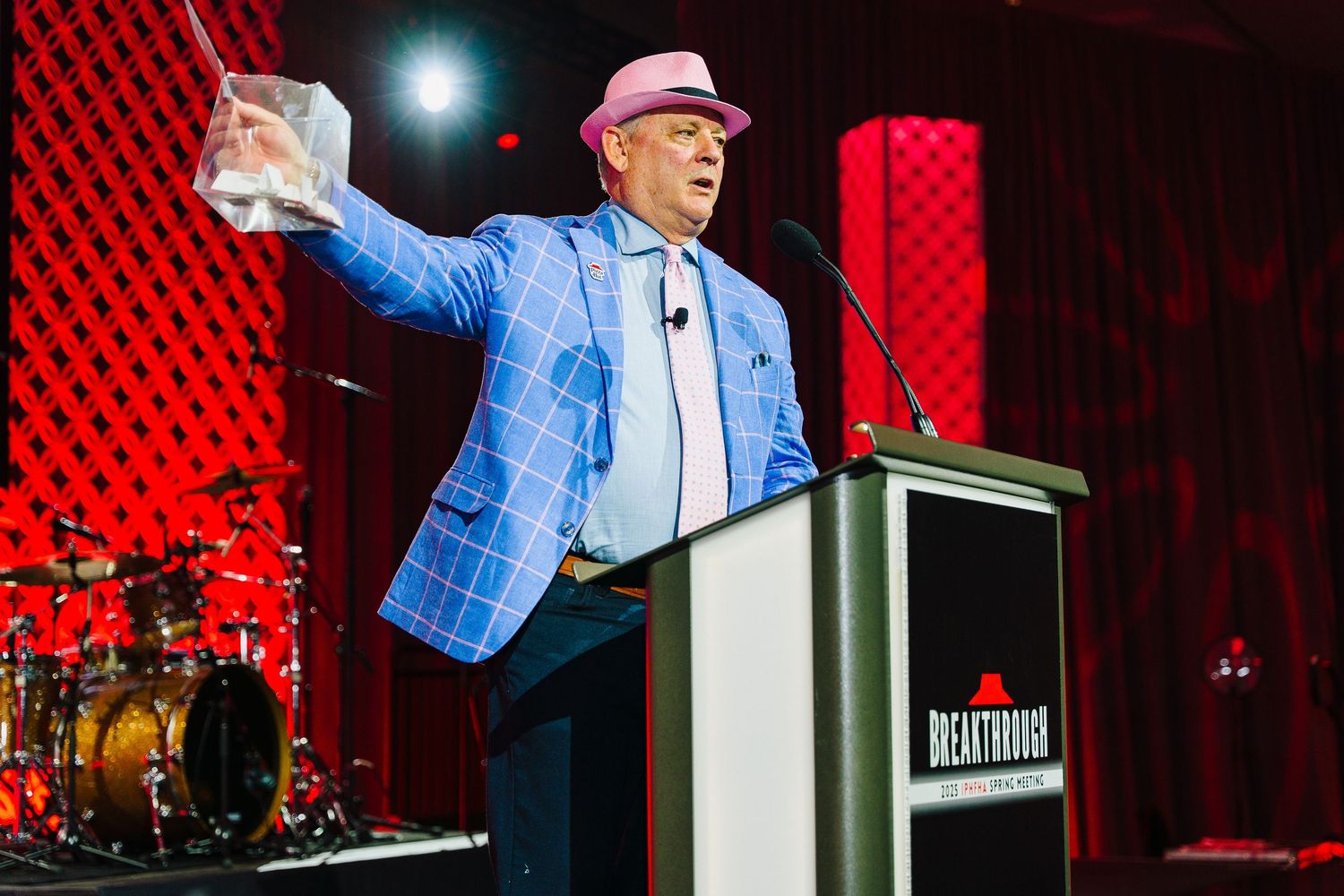 Speaker in blue blazer gesturing at podium with red backdrop.
