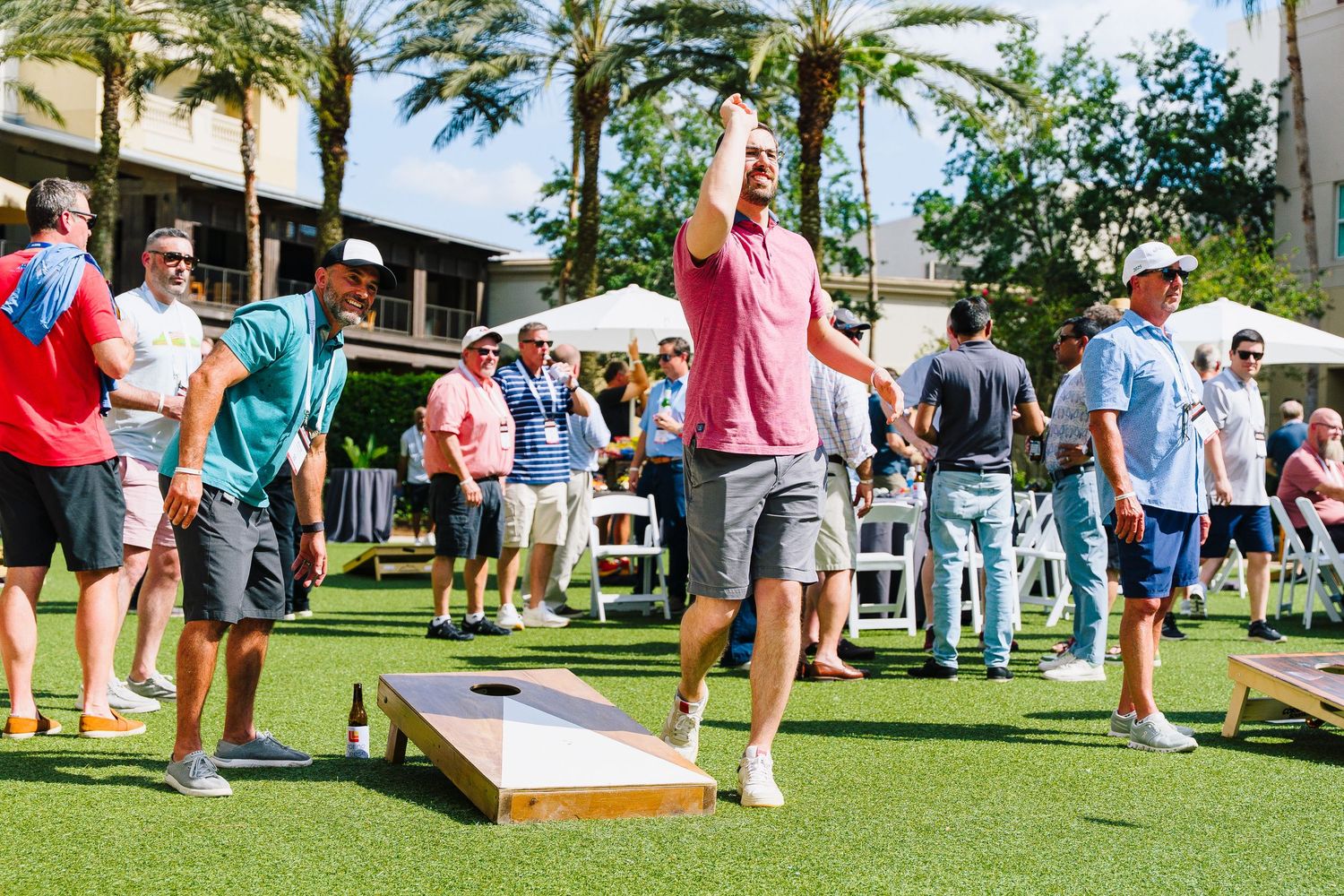 People playing cornhole game on sunny lawn with palm trees and white tents.