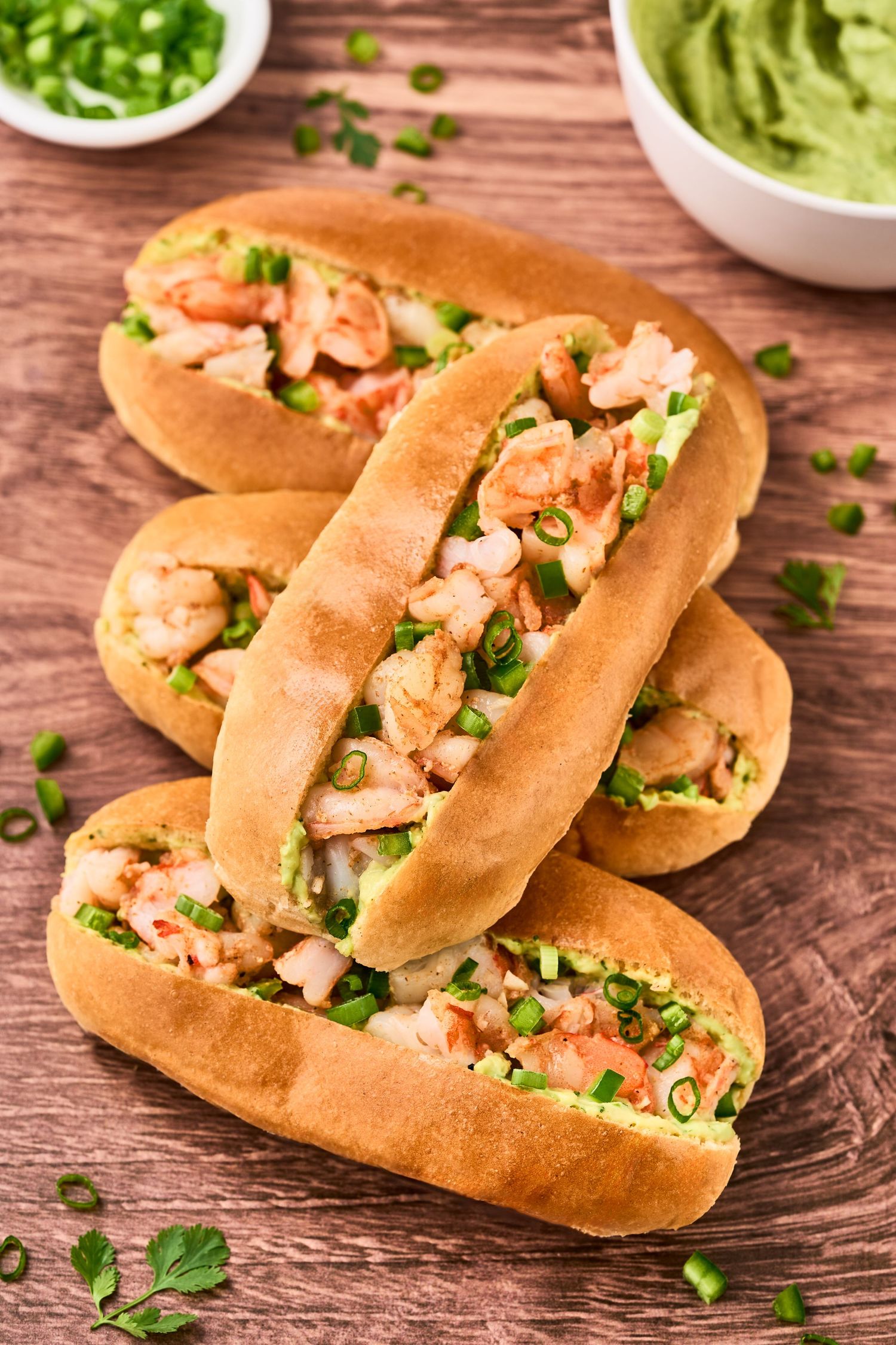 Homemade shrimp rolls on buttery toasted buns garnished with fresh herbs and served with guacamole dip on wooden table.
