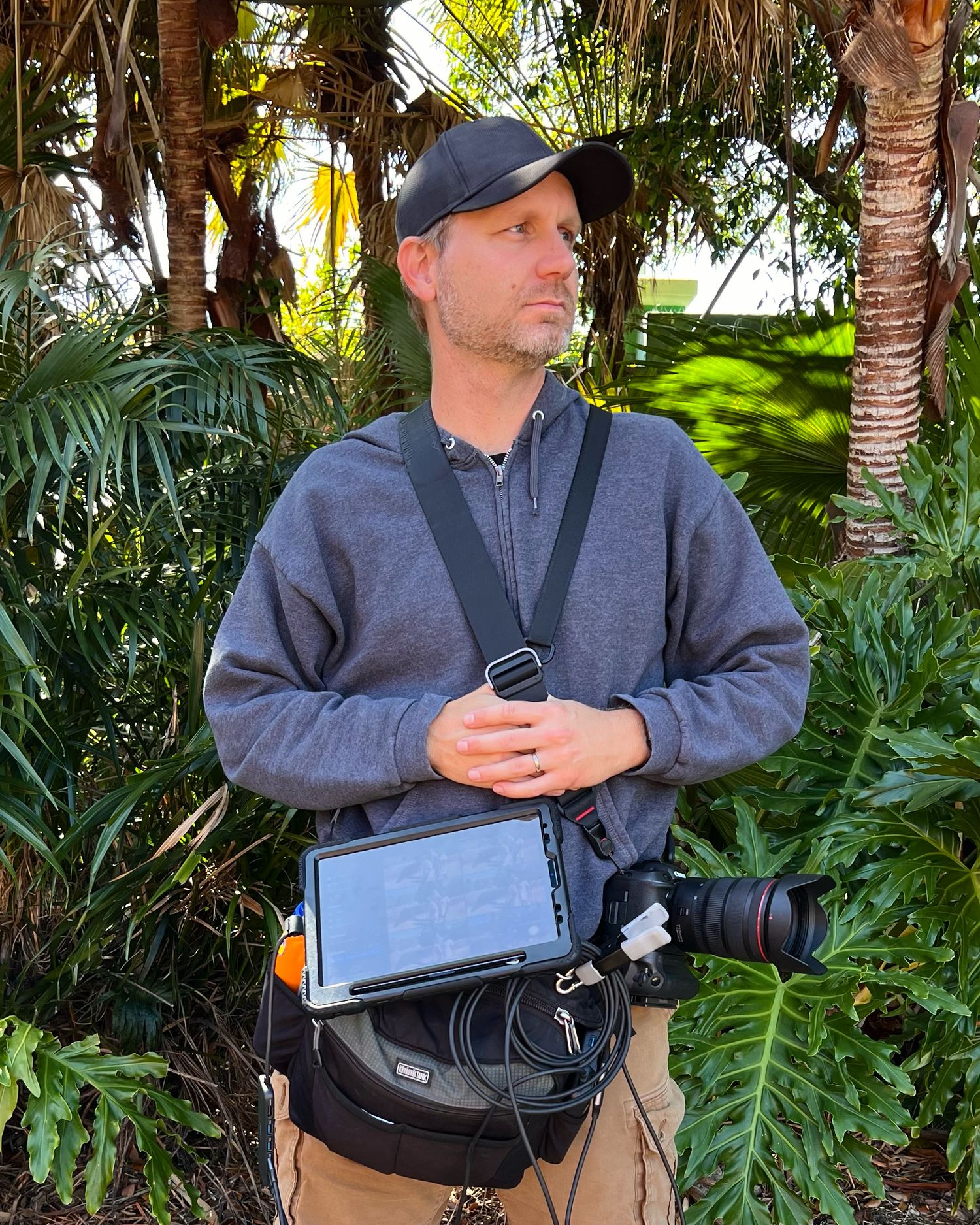 A man in a gray jacket stands against a lush tropical garden backdrop while wearing a utility bag.
