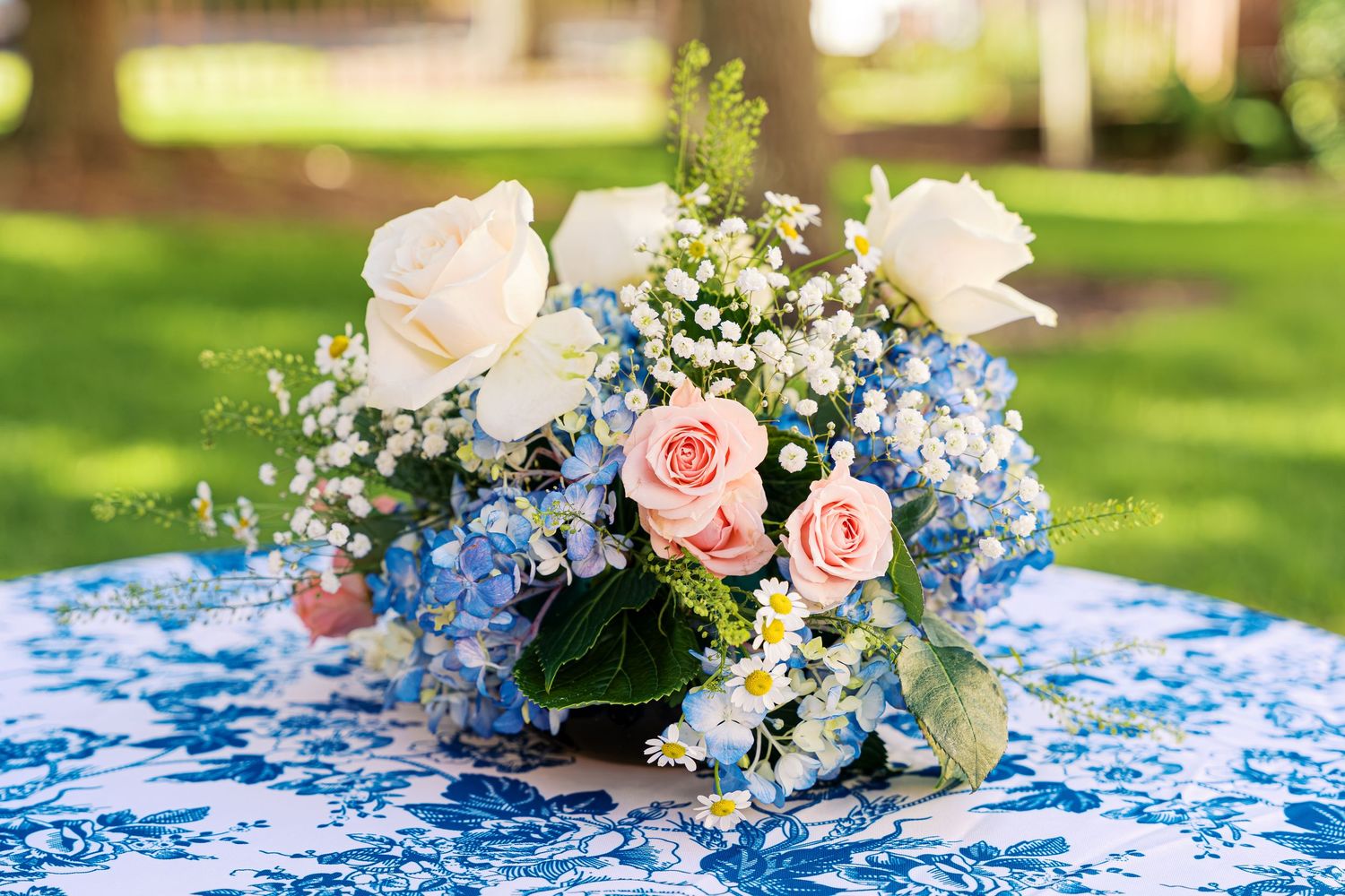 A floral centerpiece with white and pink roses mixed with blue hydrangeas sits on a blue patterned tablecloth outdoors.