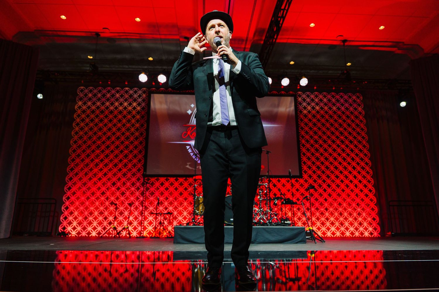 Performer in black suit energetically singing on stage with red lighting and checkered backdrop.