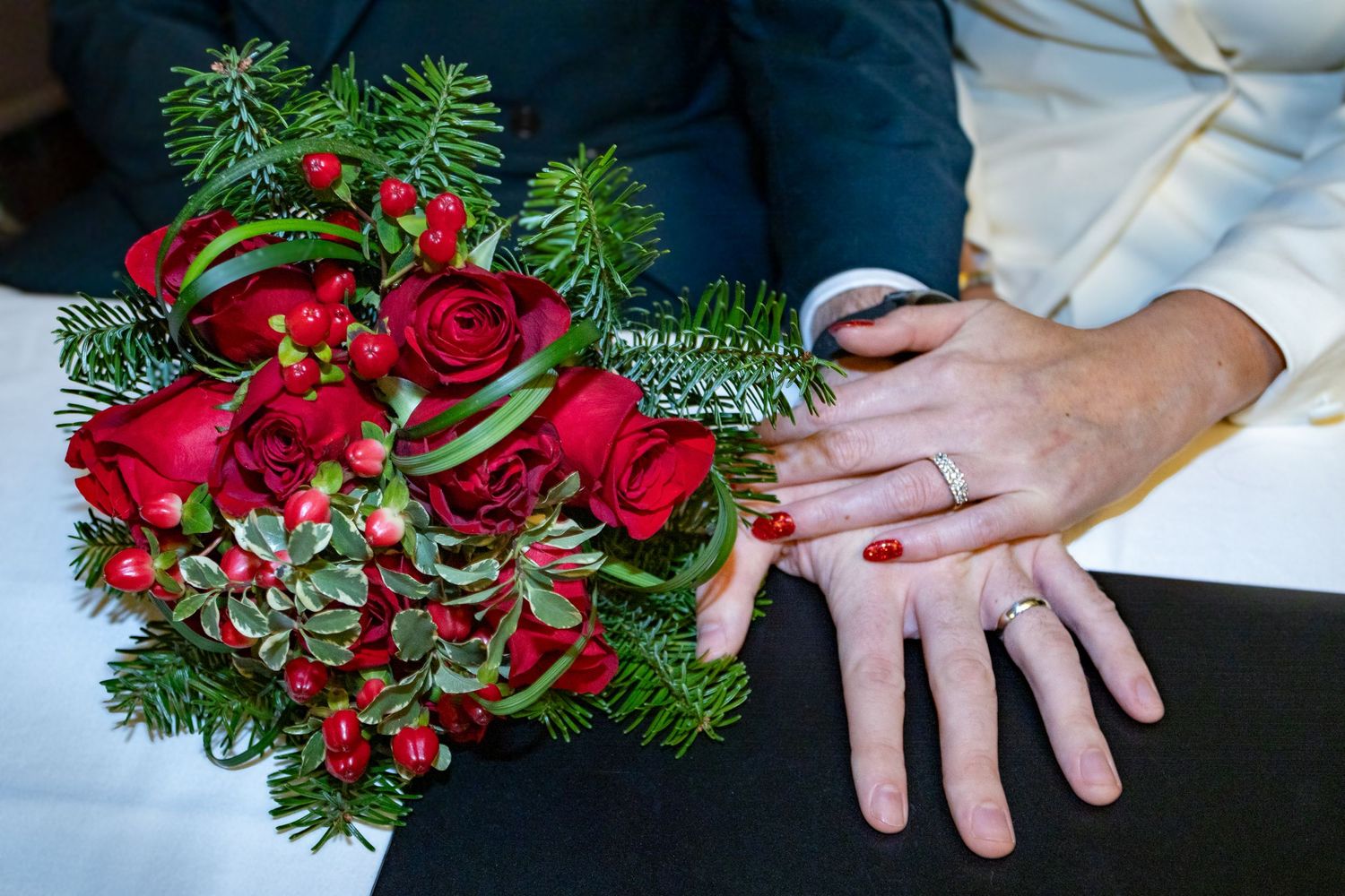 A wedding bouquet of red roses and evergreen sprigs displayed next to hands showing rings on white fabric. Wedding photo