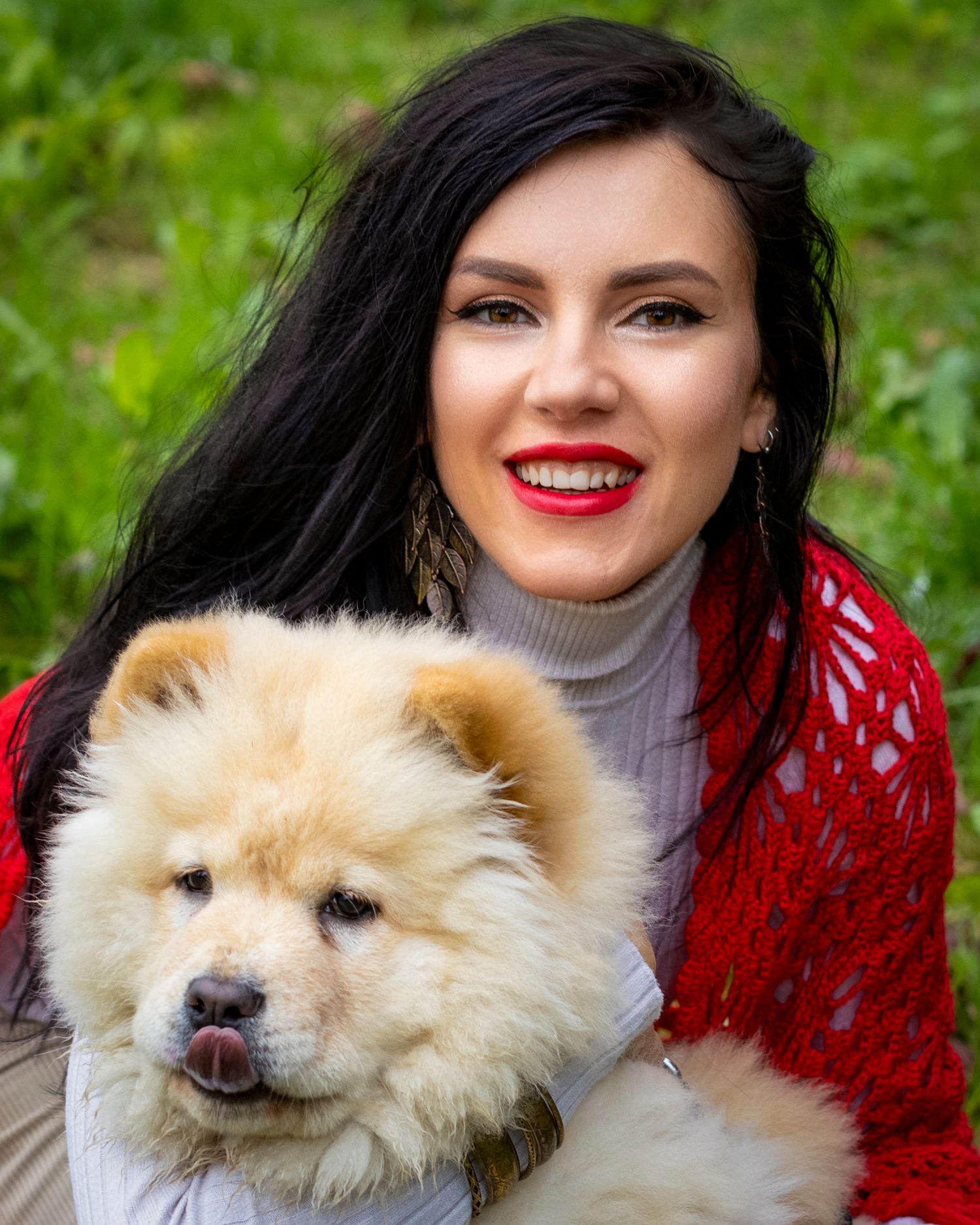 Smiling dark-haired woman in red cardigan holding a fluffy cream Chow Chow puppy outdoors in green park.