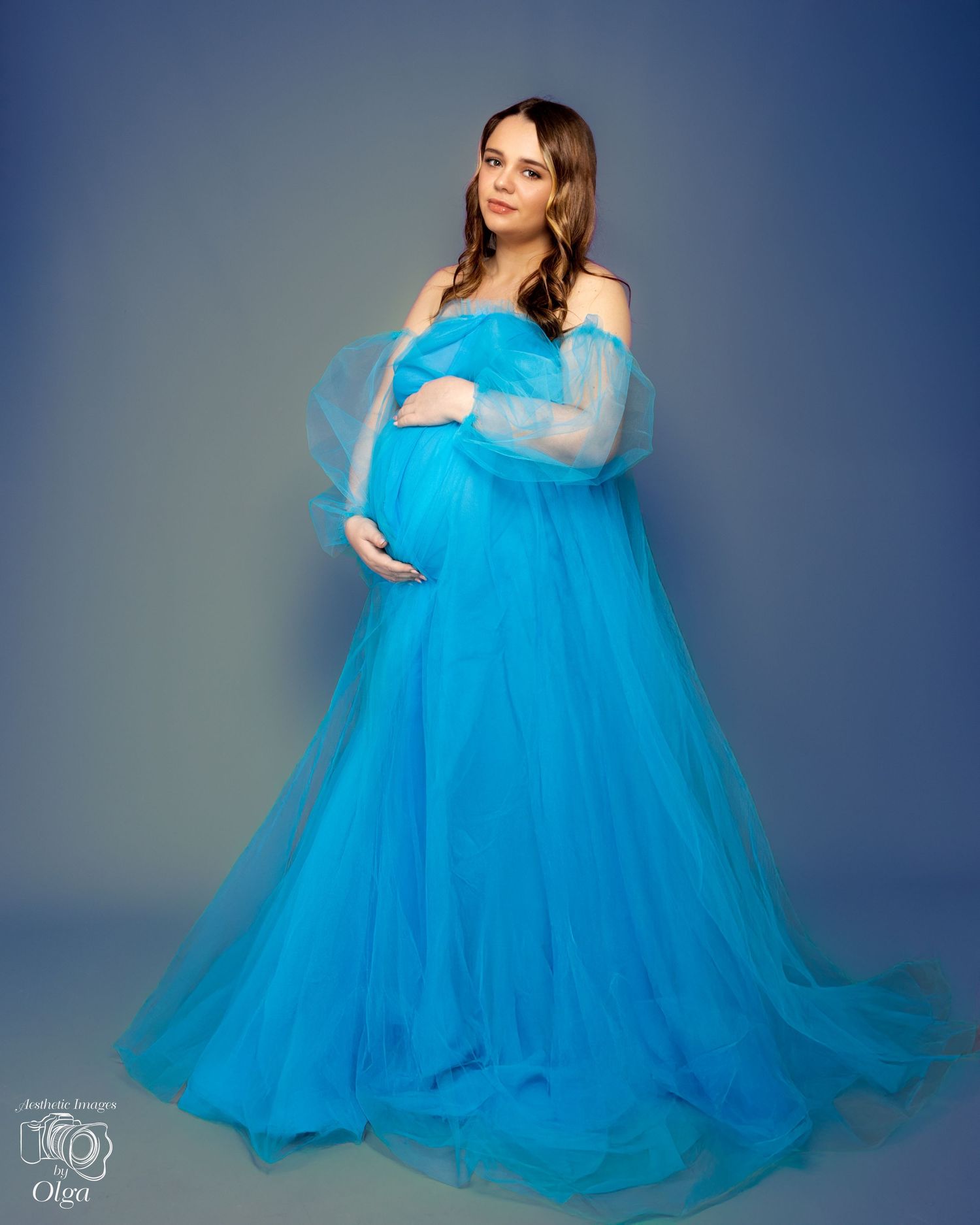 Elegant maternity session in a flowing turquoise tulle gown against a grey studio backdrop creates a dreamy aesthetic.