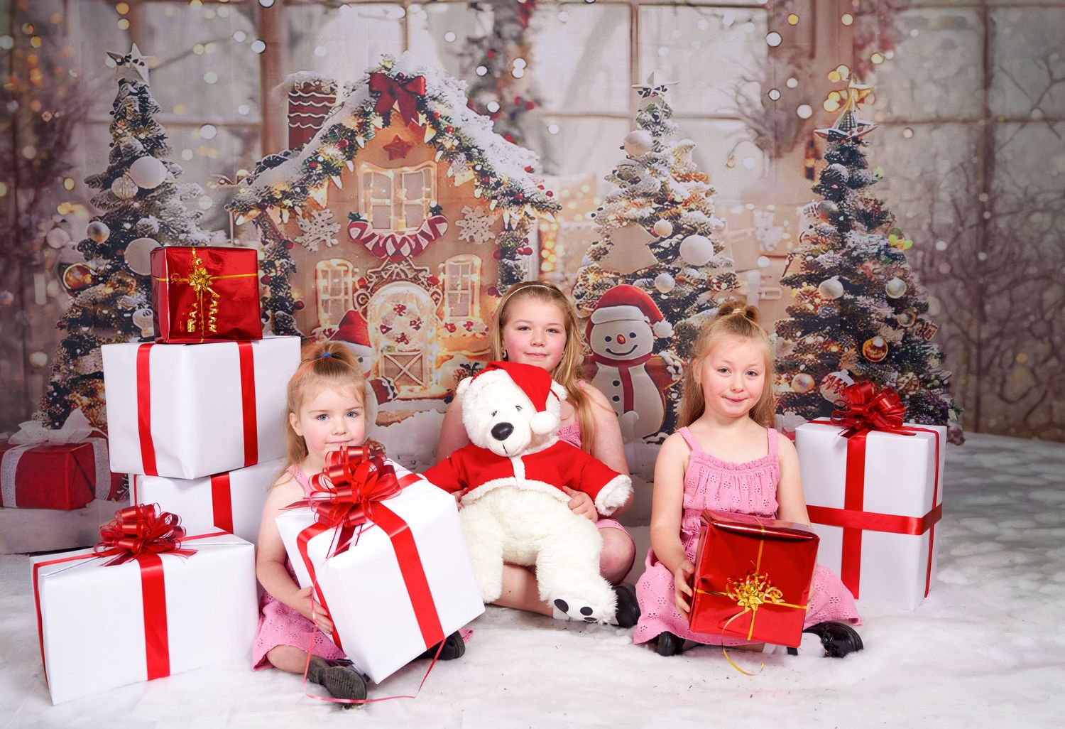 Three young girls sit on faux snow with red-ribboned Christmas presents and a teddy bear near a gingerbread house and decorated trees.