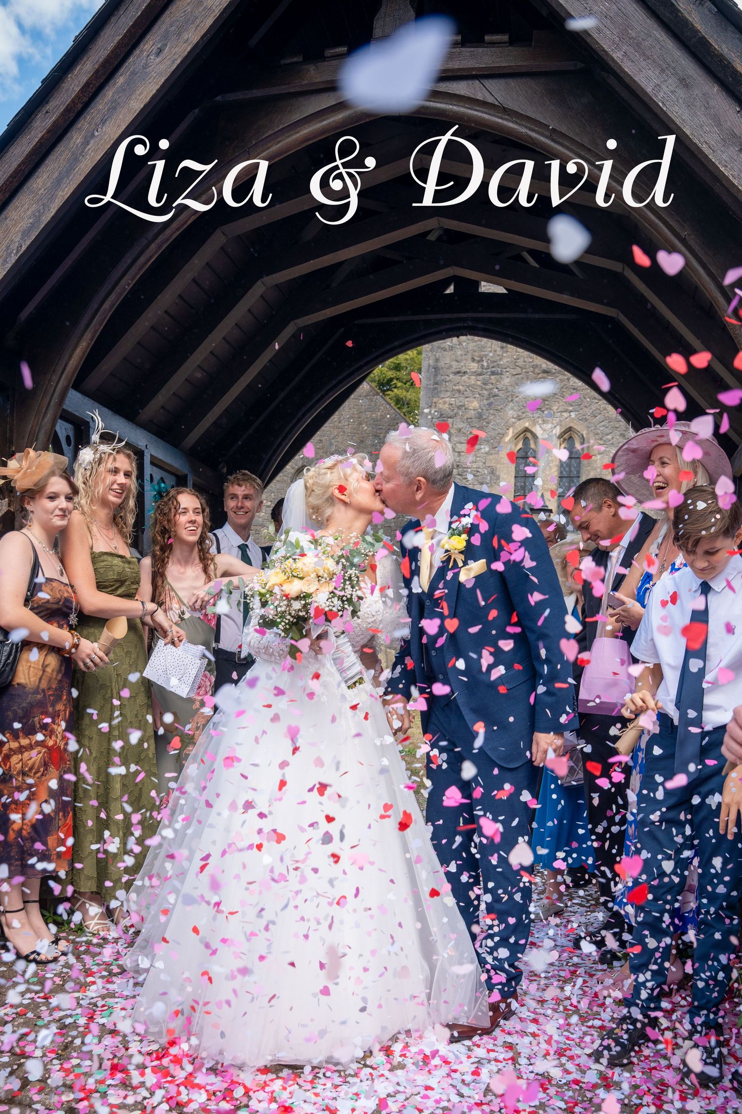Liza and David share a kiss under a church arch as guests throw colorful confetti at their wedding.