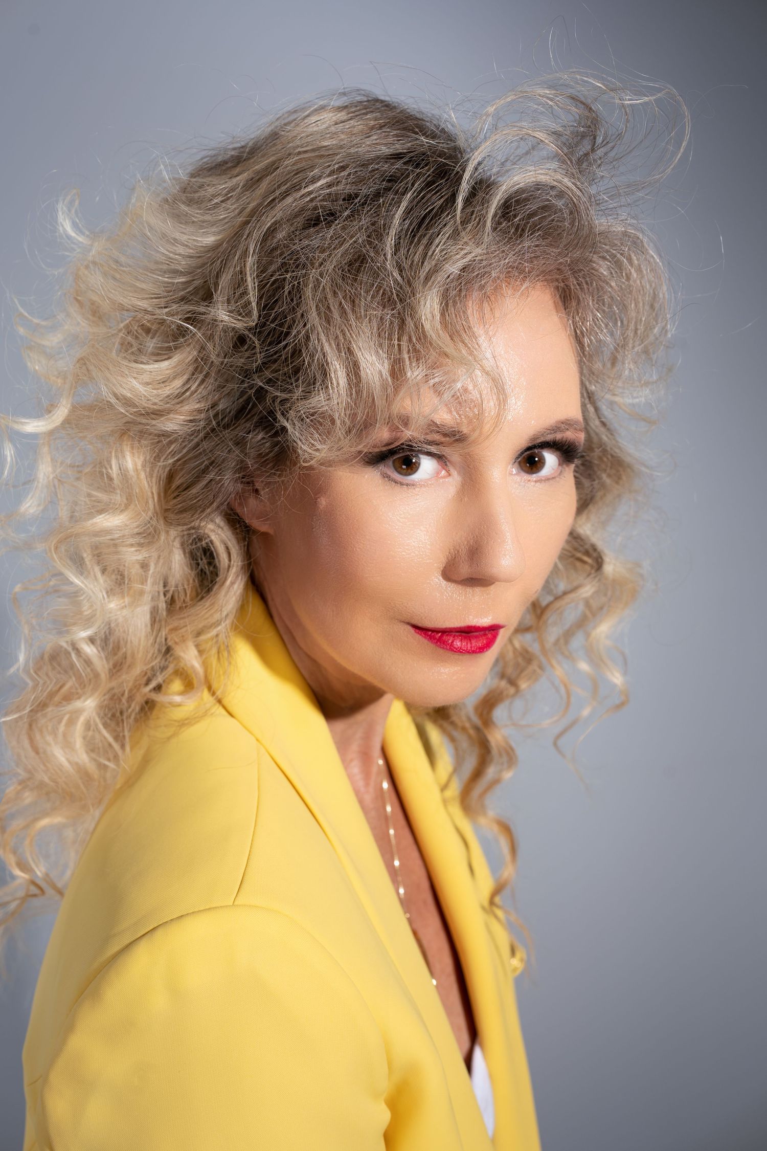 Professional portrait featuring curly blonde hair and a vibrant yellow blazer against a gray studio background.
