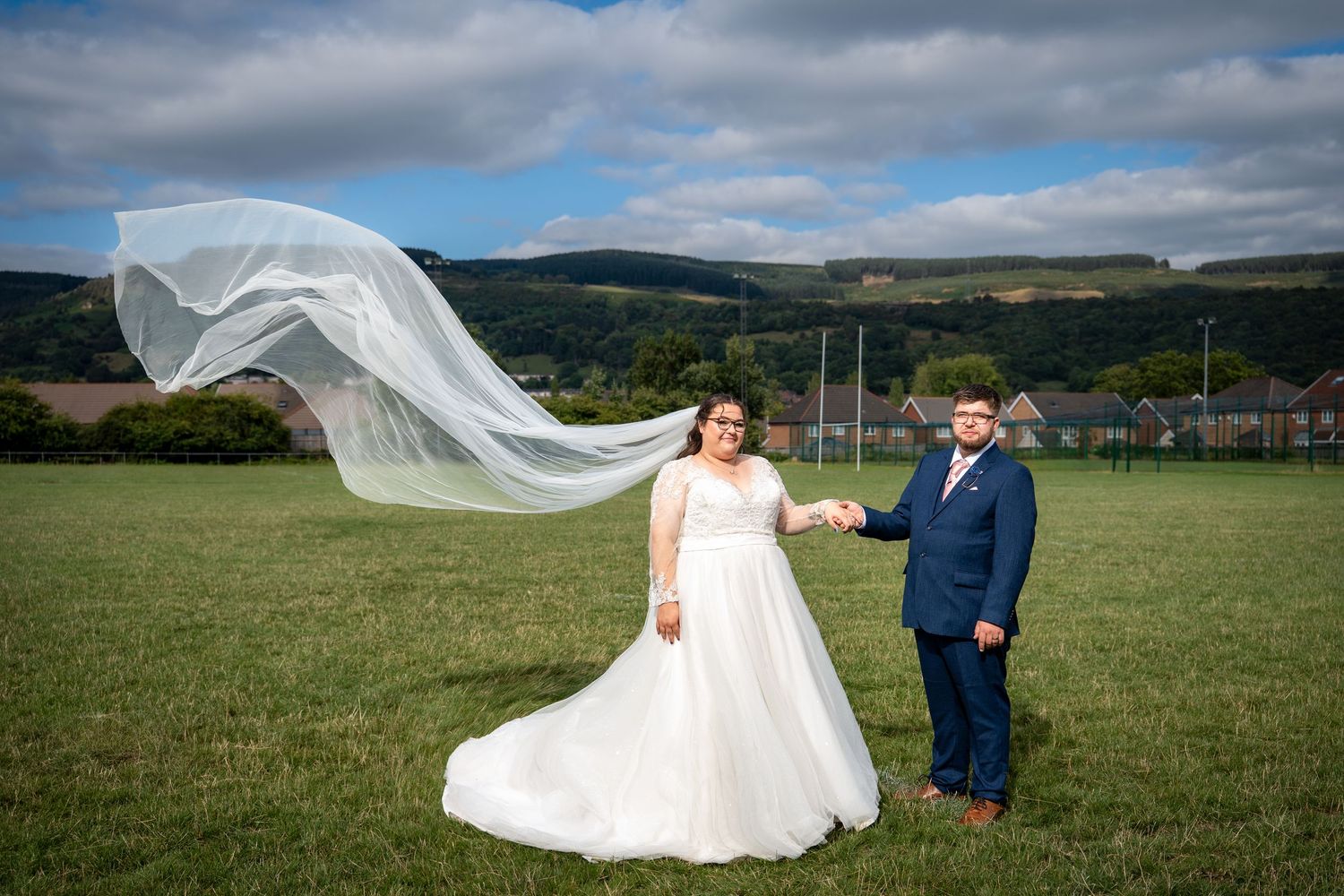 A flowing white wedding veil billows dramatically in the wind across a green field with mountains in the background.