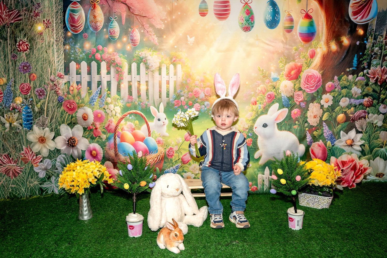 Easter backdrop display featuring colorful eggs, spring flowers, white picket fence, and bunny decorations in a garden setting.
