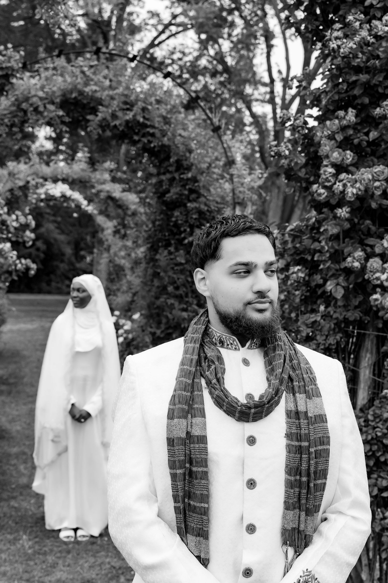 A black and white wedding photo in a garden setting with a scarf-draped person in white and a figure in white robes behind. | PHL, PA