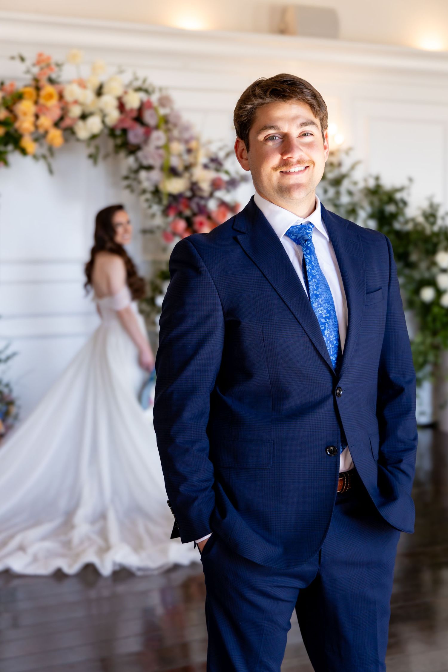 Wedding day photo shows groom in navy suit and blue patterned tie with bride's silhouette in white gown by floral arch.