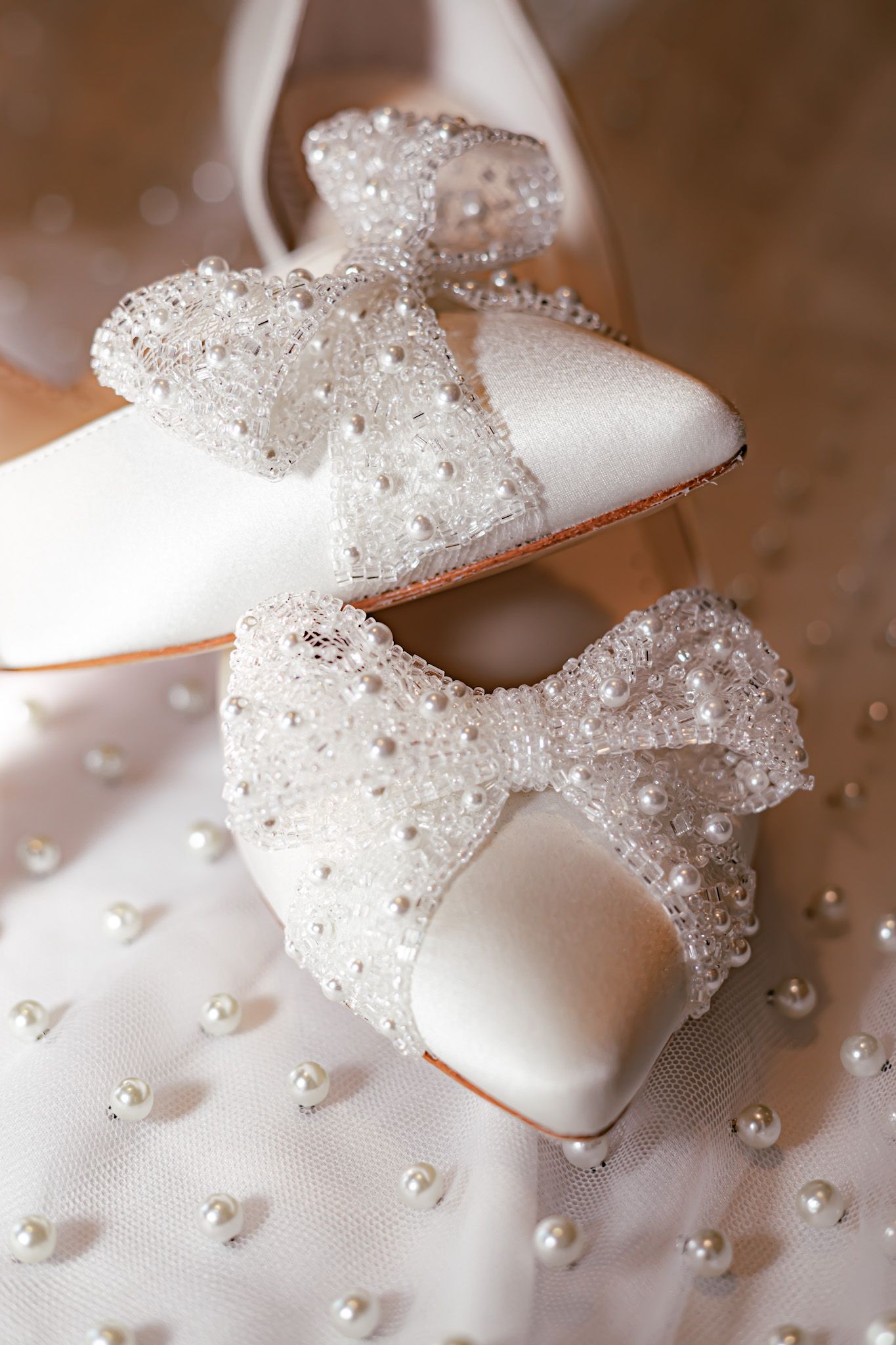 Elegant white bridal shoes with sparkling beaded bows and pearls on a textured fabric background.