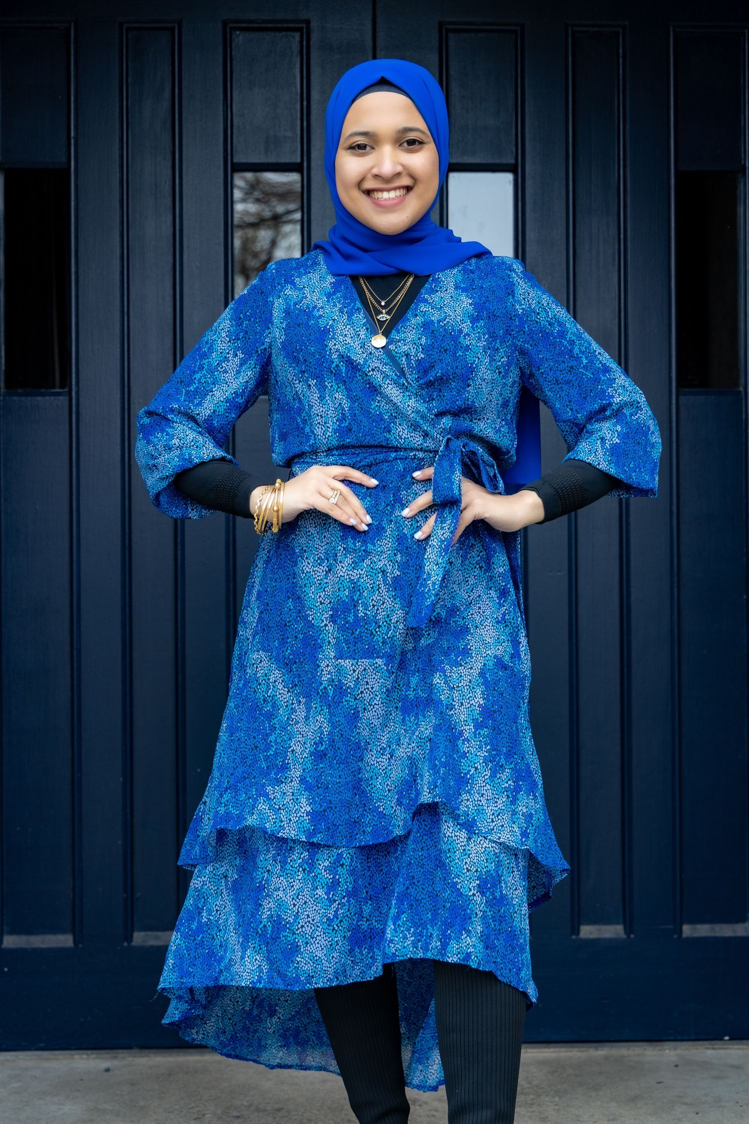 A bright blue brocade dress with layered skirt and black sleeves creates an elegant modest fashion ensemble against dark doors.